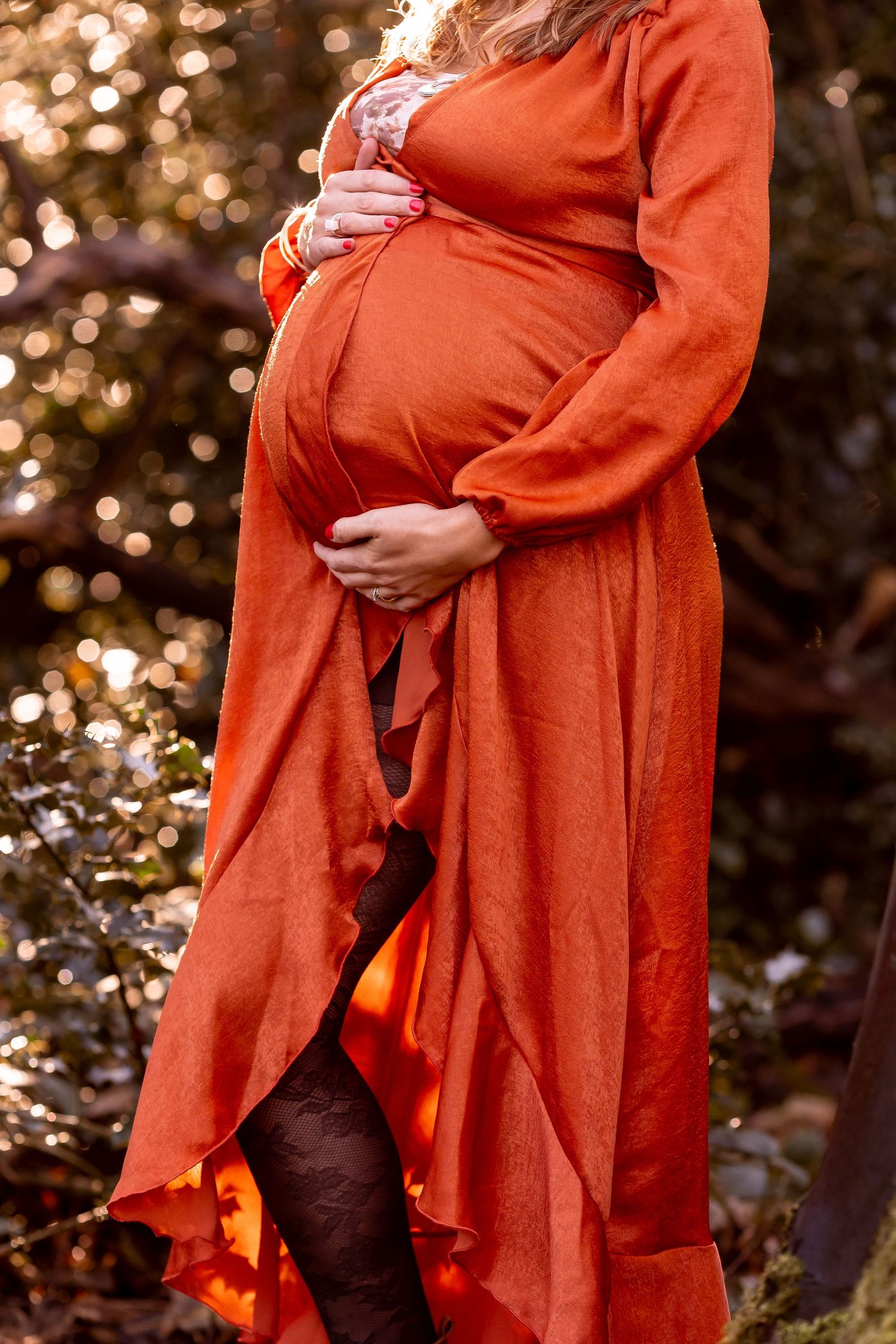 Maternity photos featuring a burnt orange dress against a bokeh forest background.