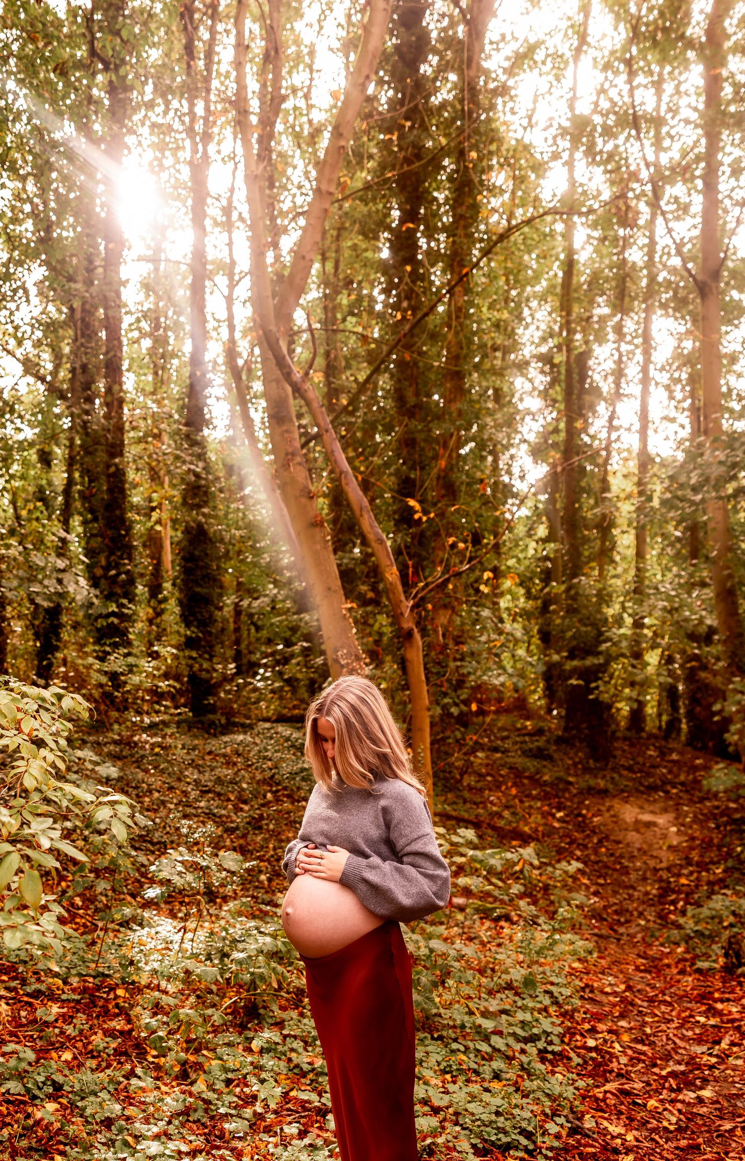 Autumn maternity photoshoot in a sunlit forest with golden leaves scattered on the ground.