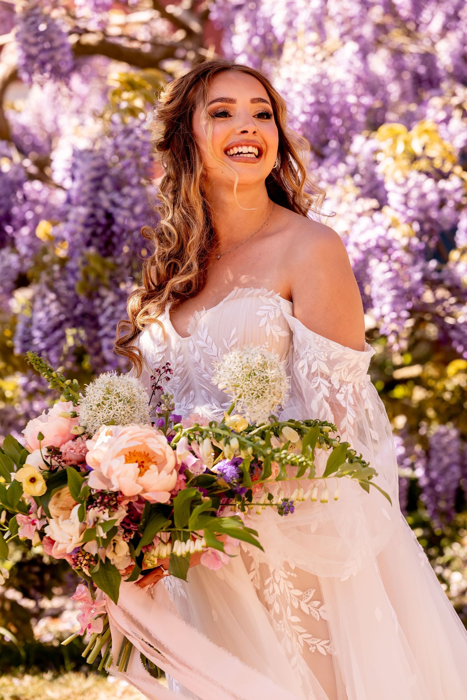 Bride in lace gown holds pink and white bouquet under cascading purple wisteria flowers.