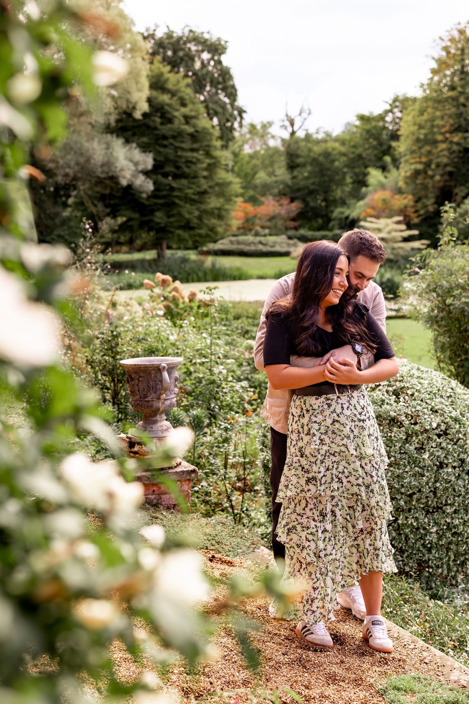 A romantic couple embraces in a lush garden setting surrounded by blooming flowers and greenery.