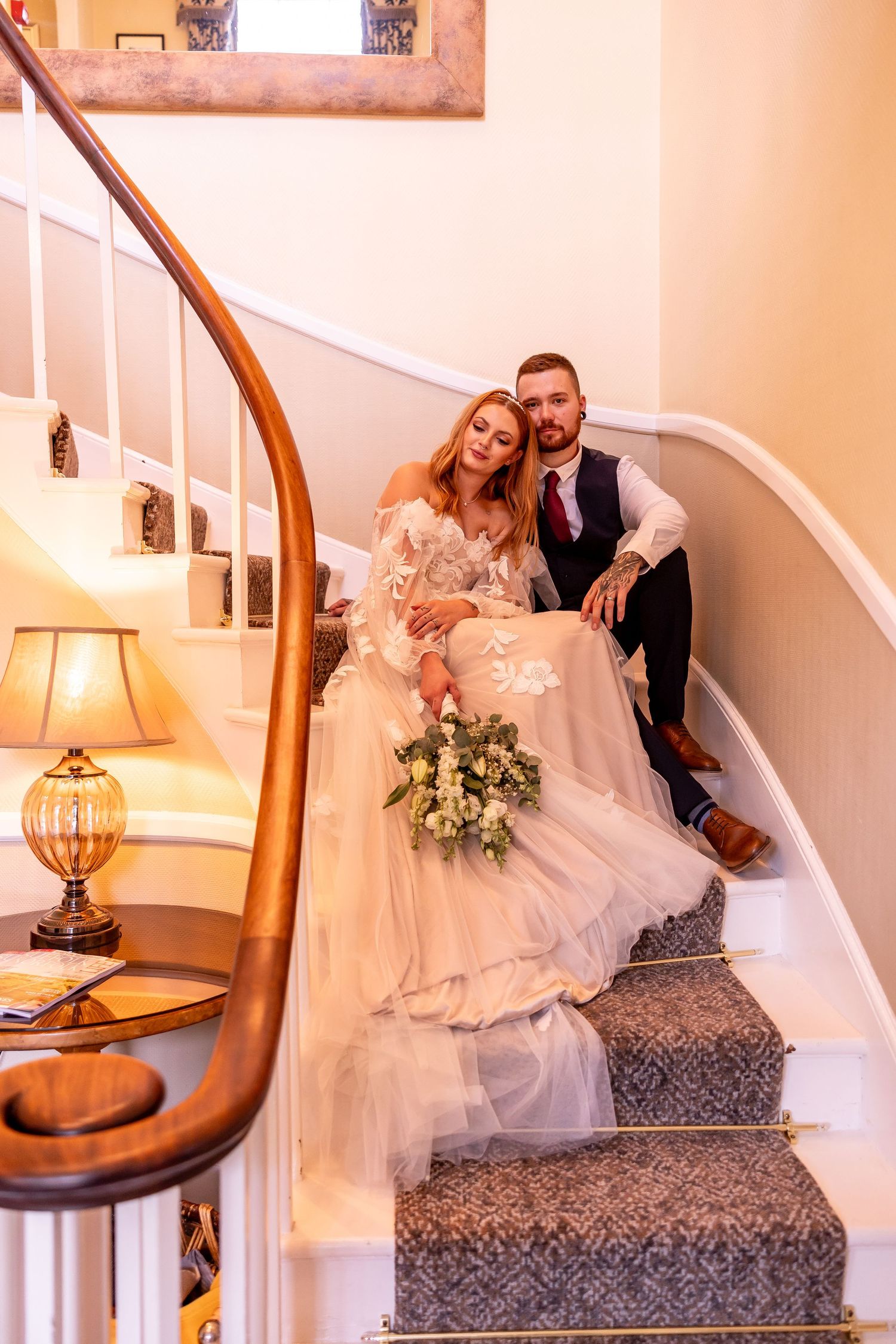 Newlyweds pose on elegant carpeted staircase with vintage lamp and flowing wedding dress.