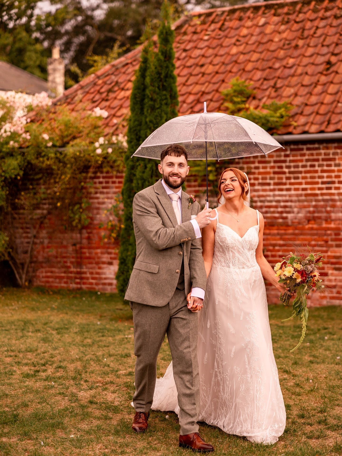 Wedding couple walking and laughing while sharing an umbrella in front of a brick building.