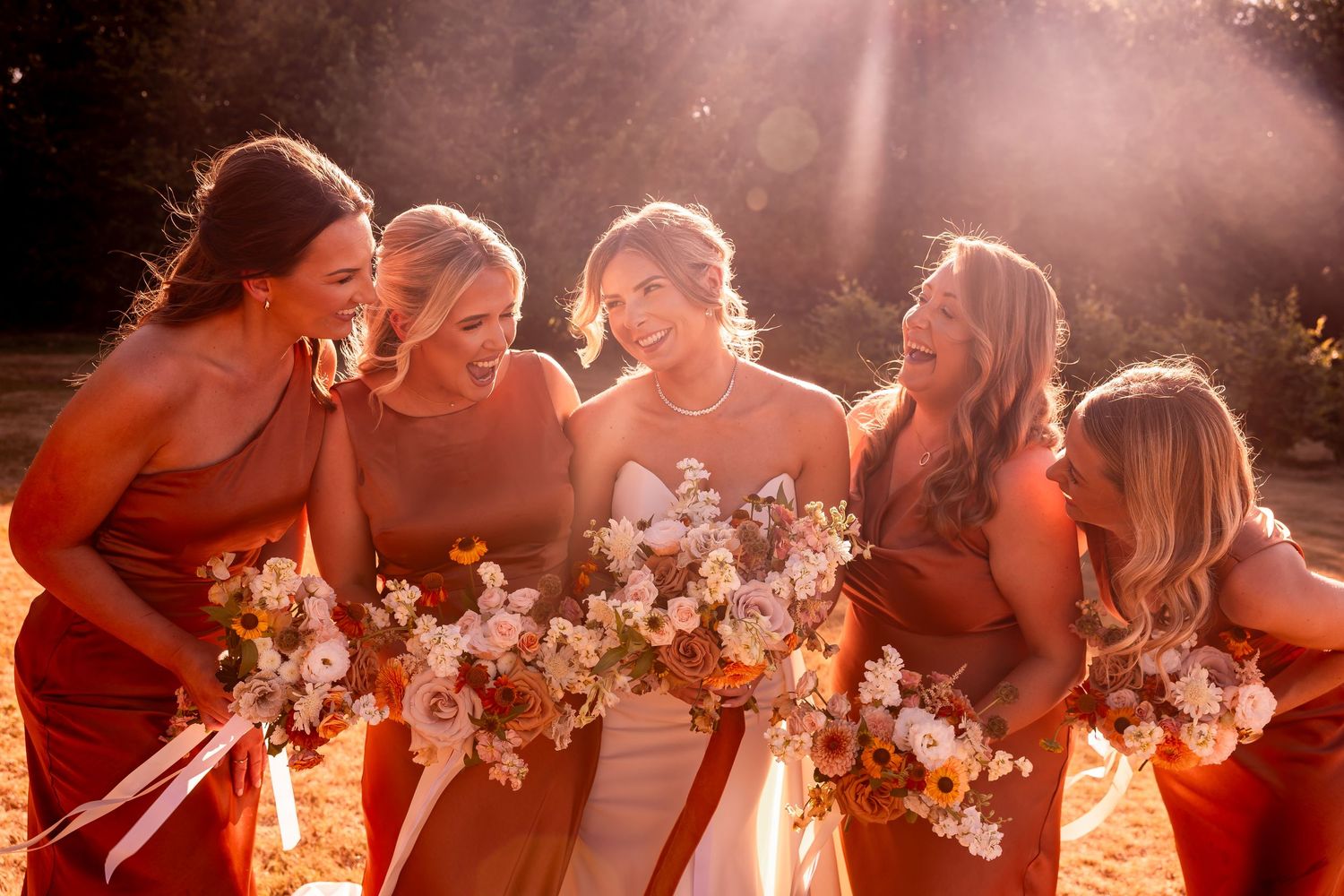 Bridesmaids in rust-colored dresses share a joyful moment while holding white floral bouquets in golden sunlight.