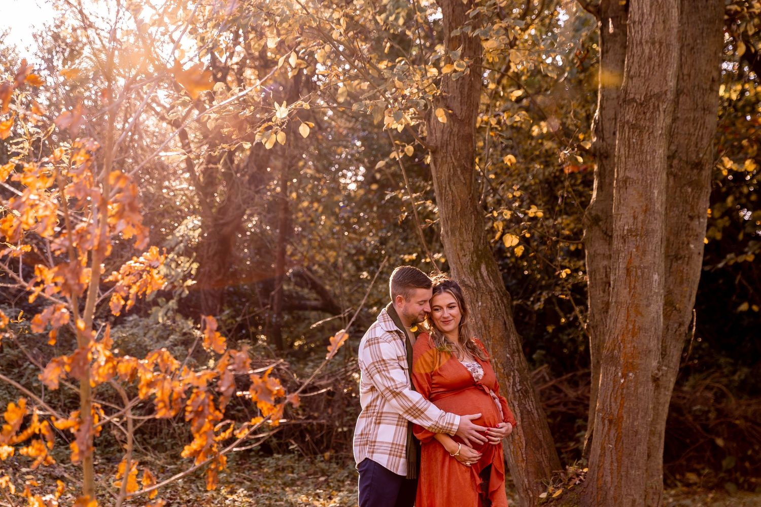 Autumn-themed maternity photos captured in a wooded area with warm golden lighting.