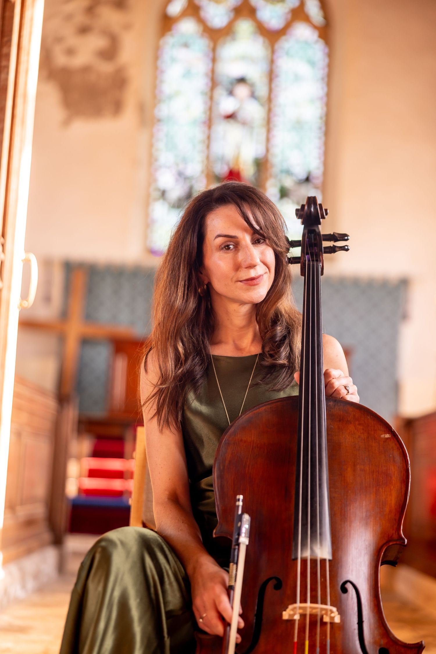 Cellist in olive green dress performs in church interior with stained glass windows.