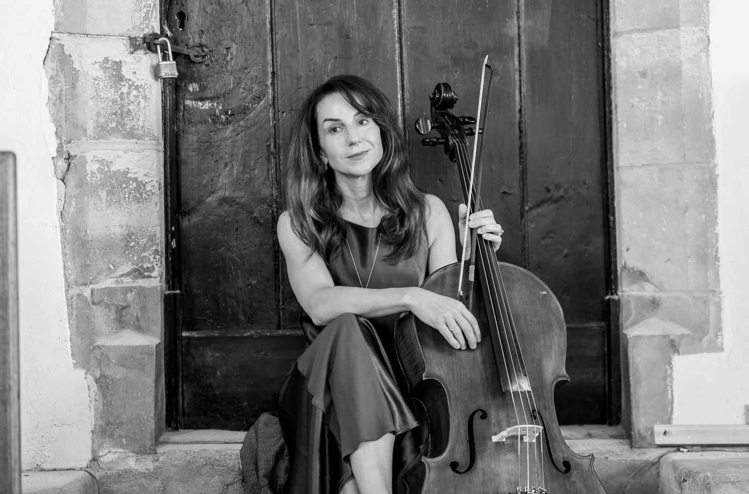 Musician with cello poses against rustic black doors in black and white.