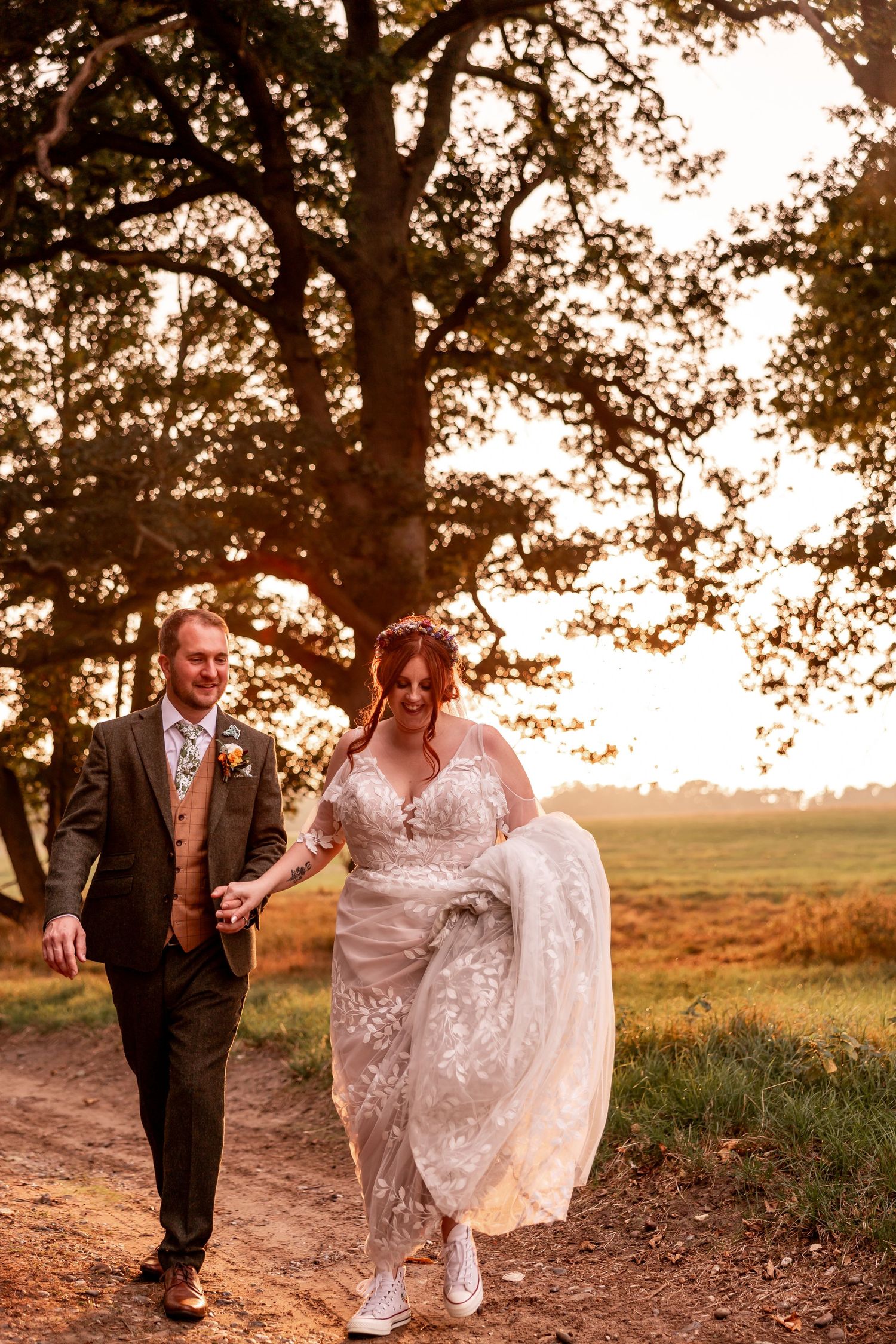 Newlyweds walk hand in hand at sunset under large oak trees in countryside setting.