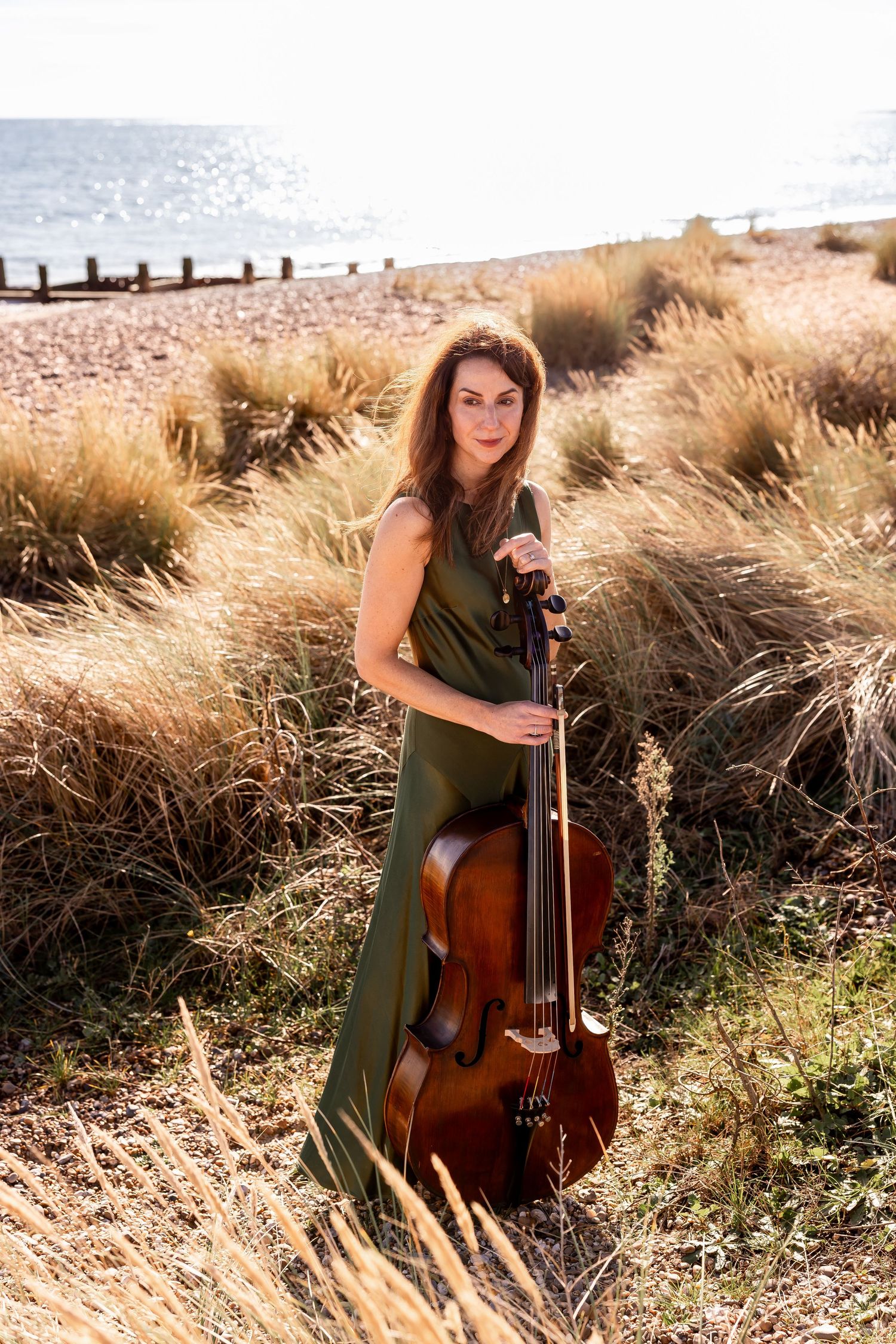 Cellist in green dress poses with instrument among tall coastal grasses at sunset.