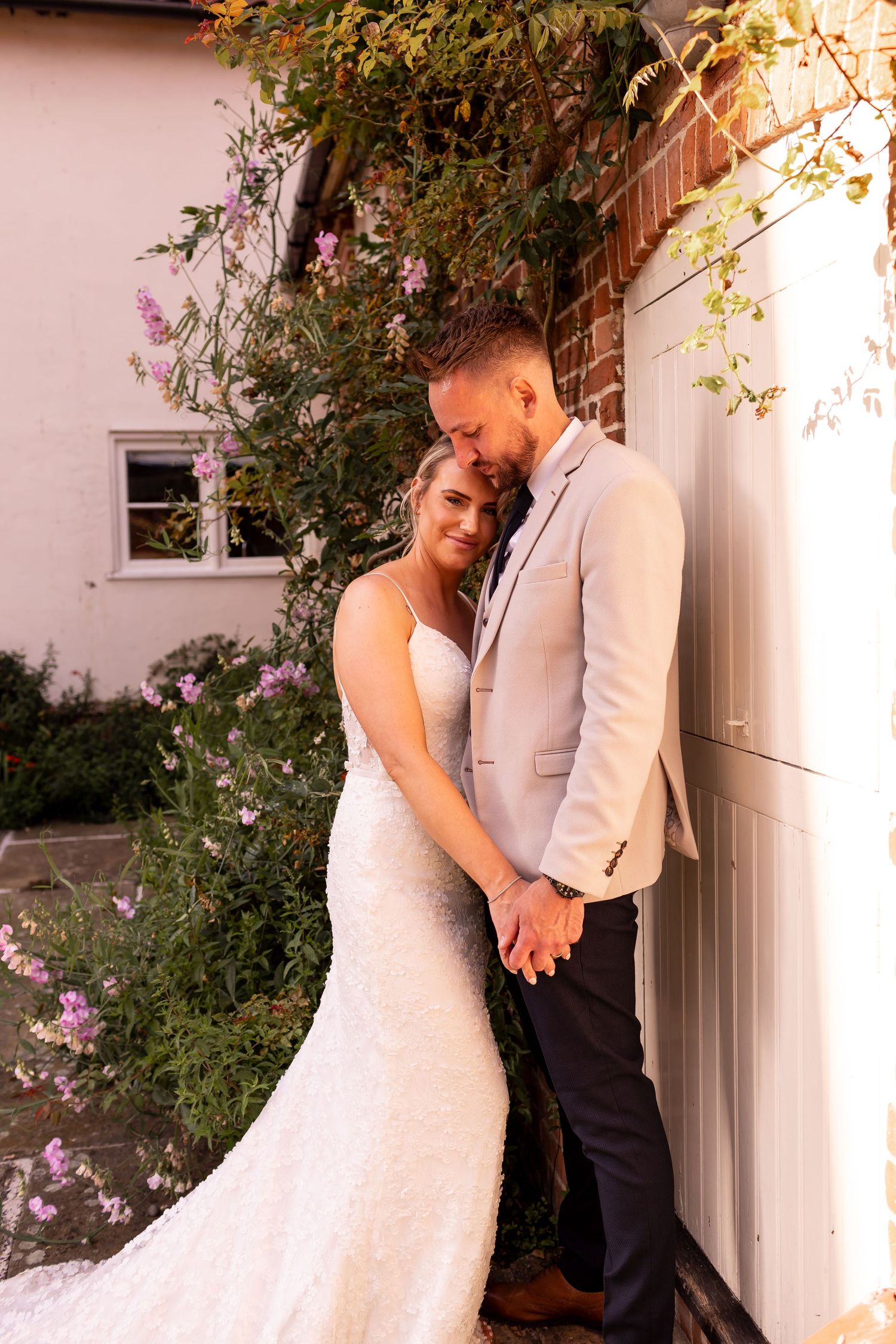 A romantic wedding couple embraces against a white wall with flowering vines in an outdoor garden setting.