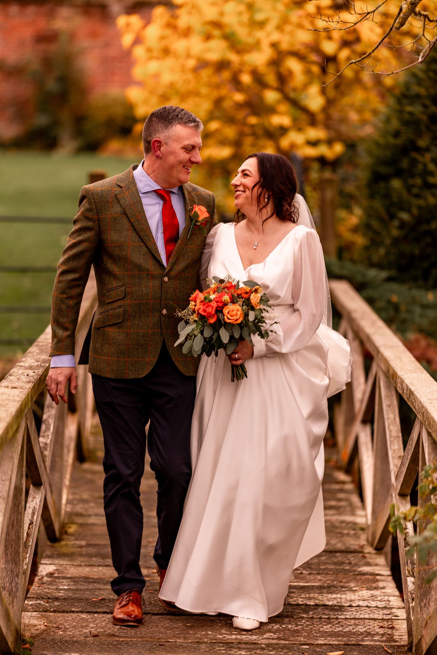 A couple shares a laugh while walking across a wooden bridge surrounded by autumn foliage.