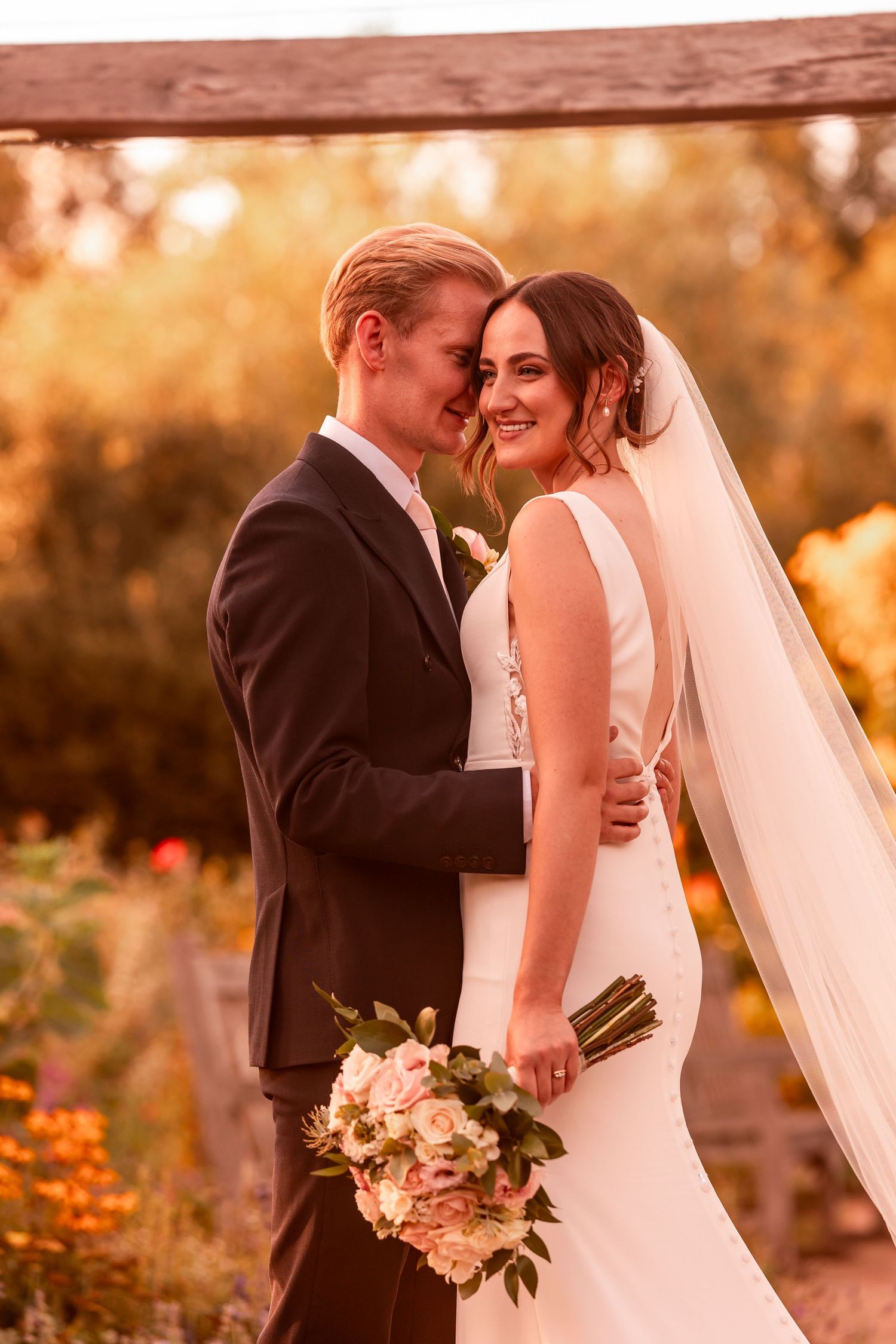 Newlyweds share an intimate moment in golden sunset light with bride holding a soft pastel bouquet.