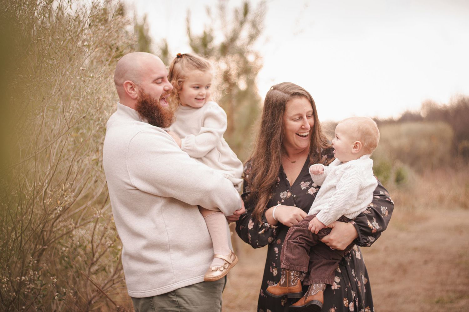 Happy family of four posing outdoors in a natural setting, parents holding two young children while smiling and laughing.