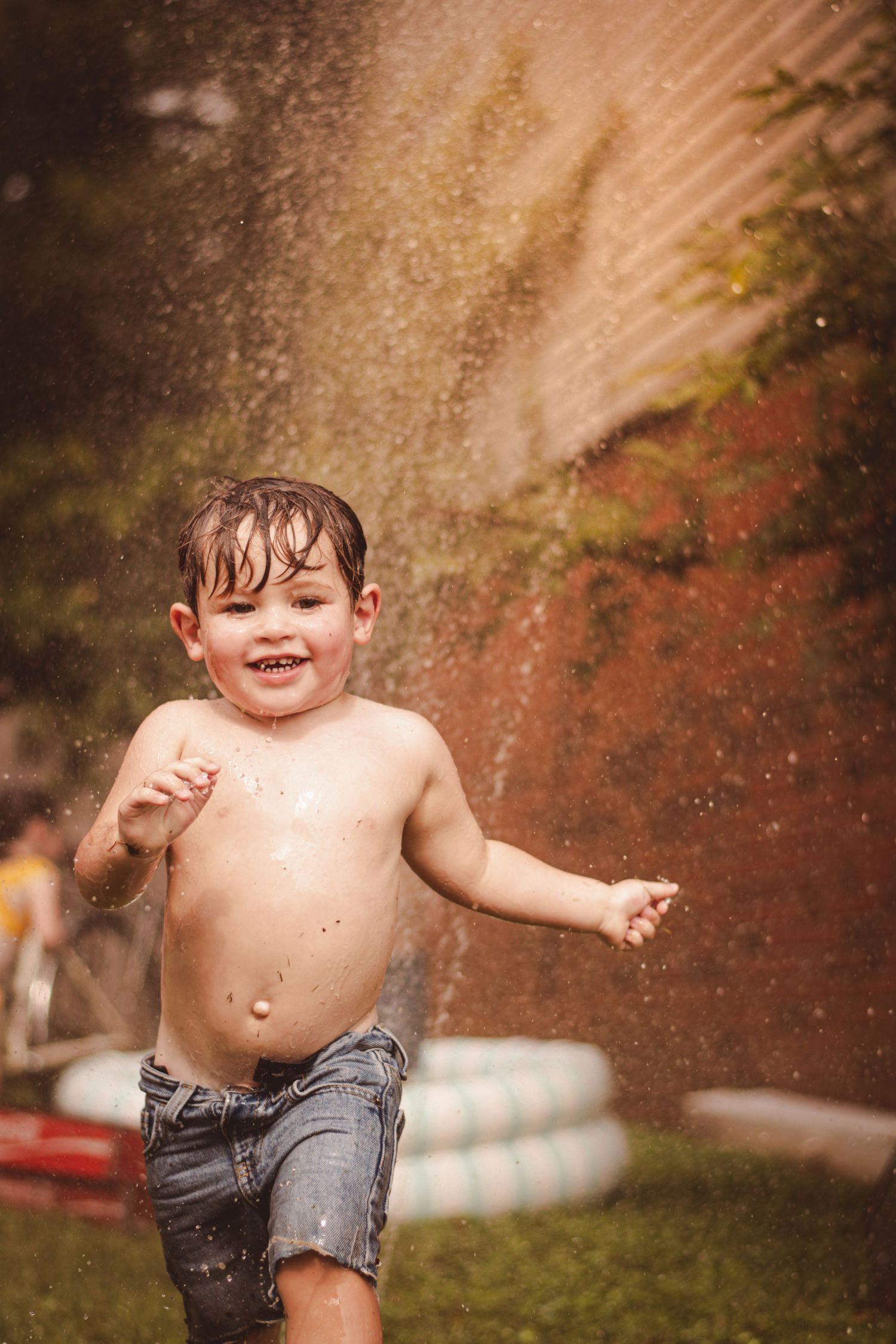 A joyful toddler boy playing in backyard sprinkler water, shirtless in denim shorts, laughing with delight on a summer day.