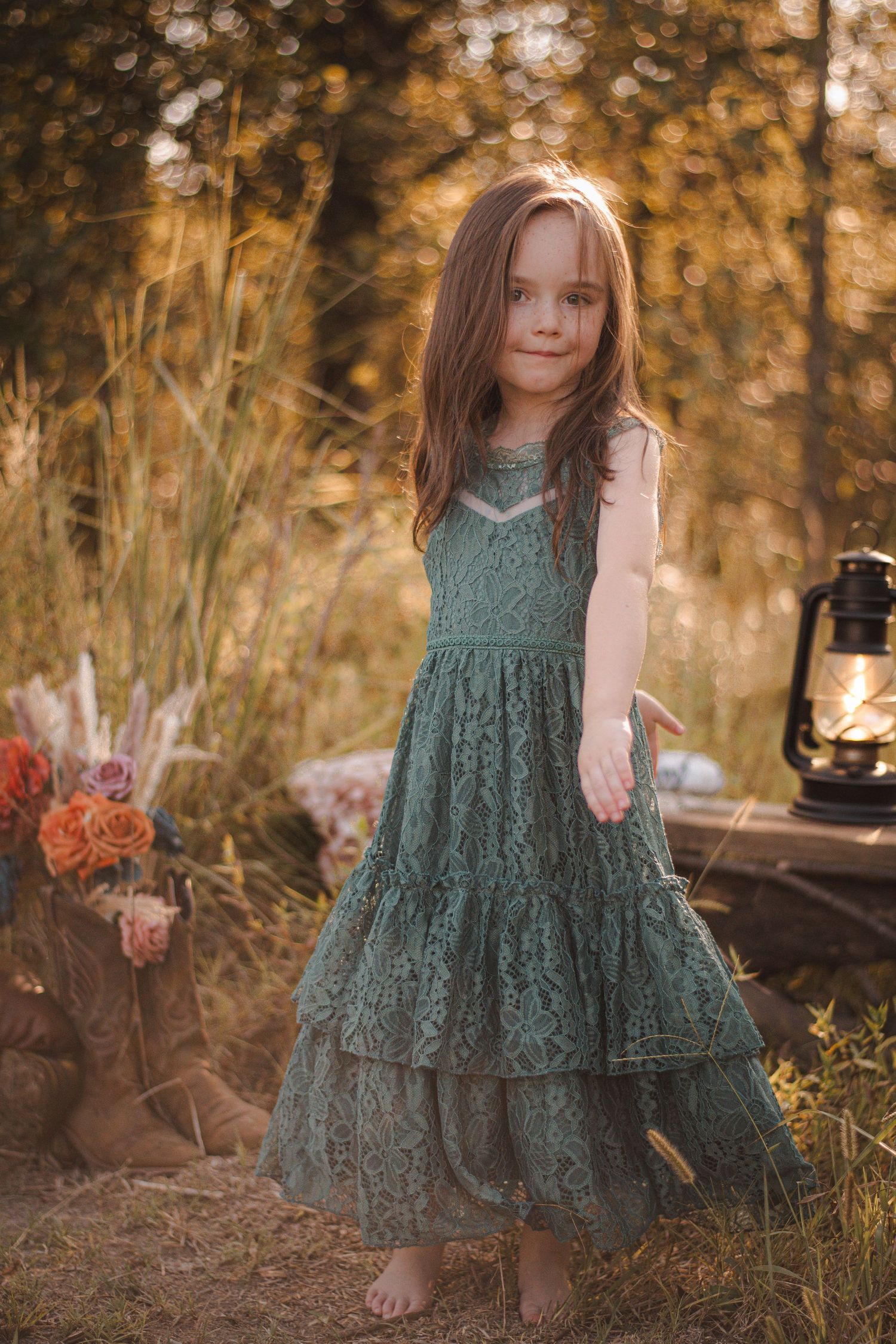 Young girl in green lace dress poses outdoors at golden hour with flowers and lantern.