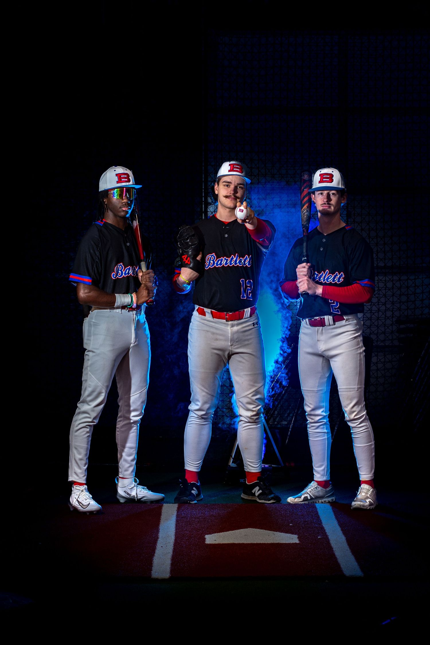 Three Bartlett baseball players posing at home plate in dramatic blue-lit stadium setting.