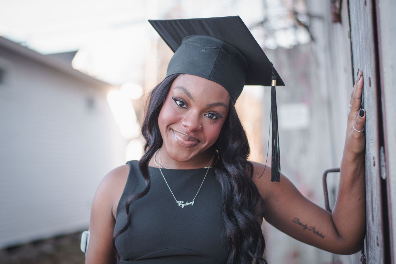 Young woman in black graduation cap and dress smiling confidently outdoors during graduation photo session.