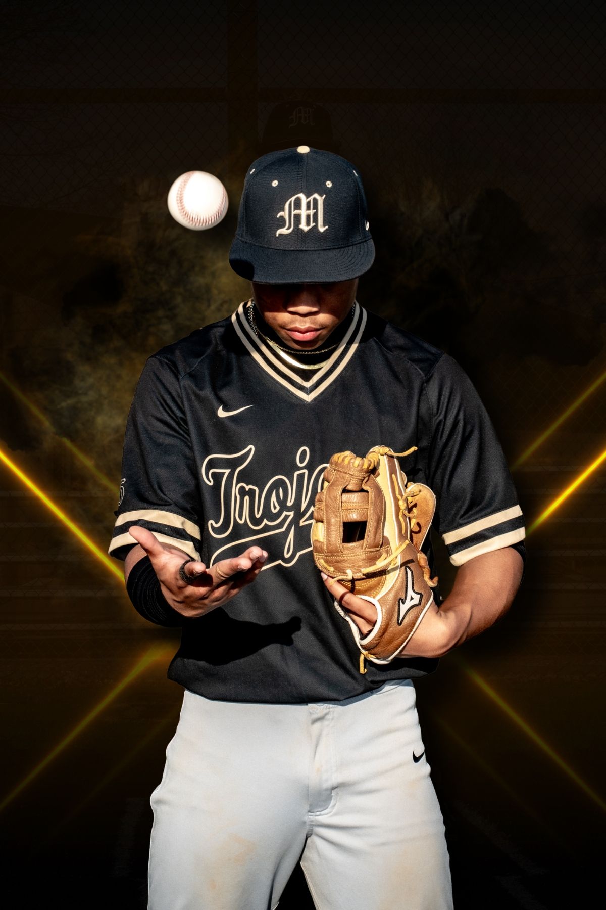 Baseball player in Trojans uniform tossing a ball while holding a glove under dramatic golden lighting.