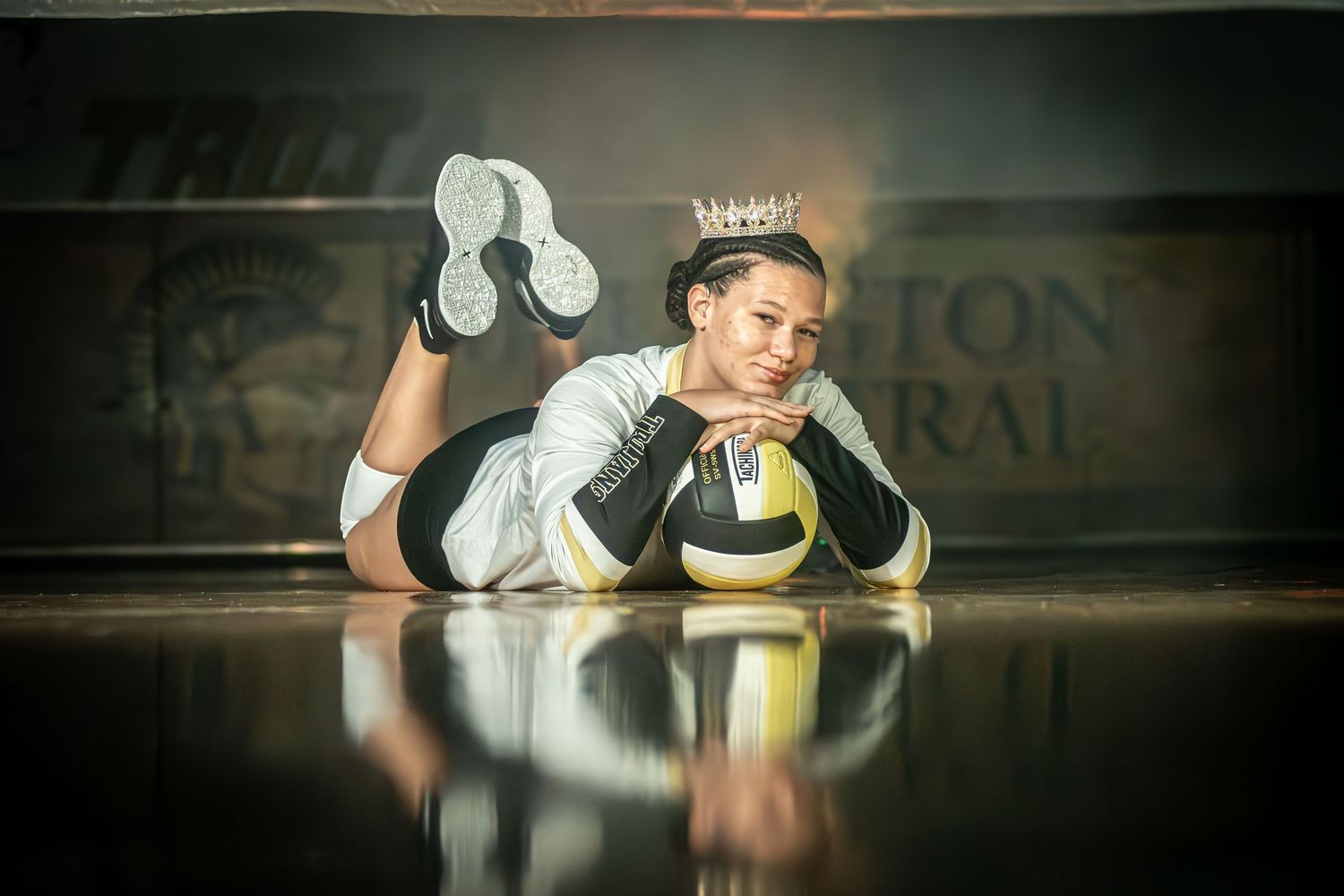 Volleyball player lying on court floor wearing a crown with a volleyball, smiling at camera.