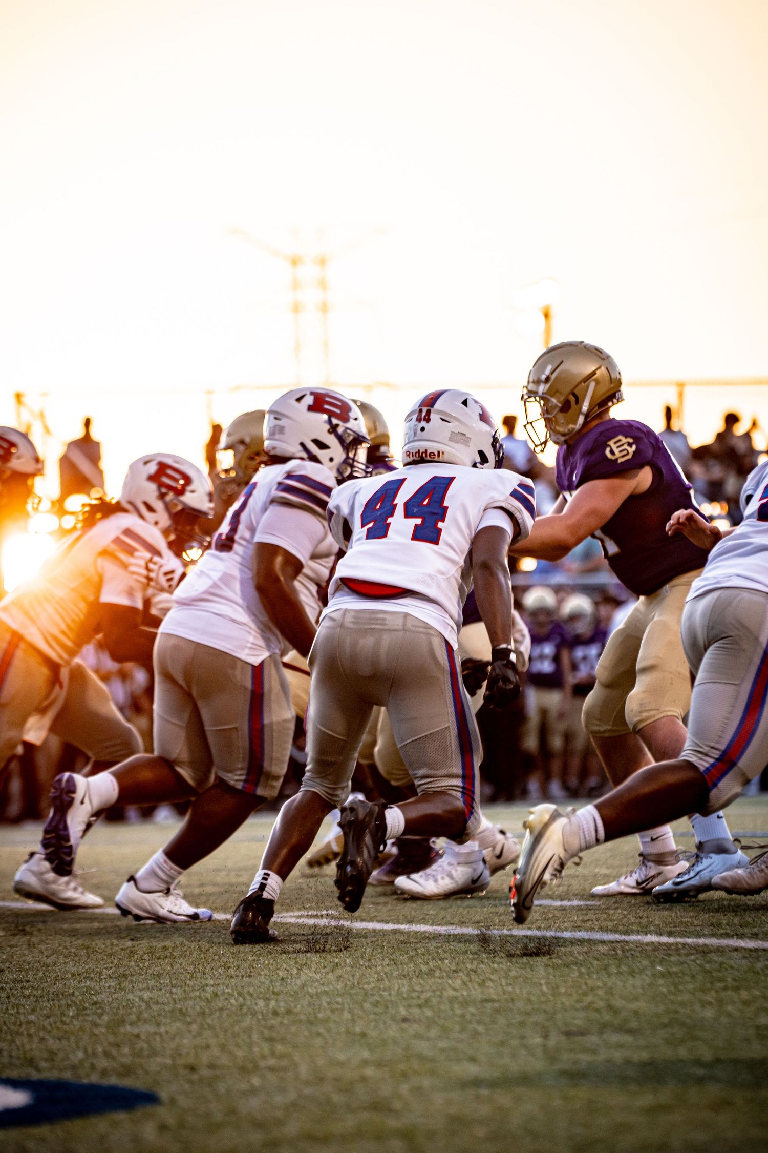 Football players clash at the line of scrimmage during a sunset game, with player #44 in white leading the defense.