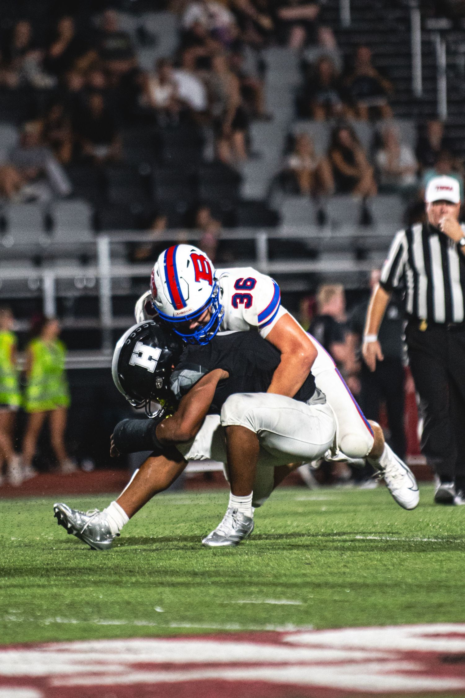 Football player #36 in white uniform tackles opponent during night game with referee and crowd visible.