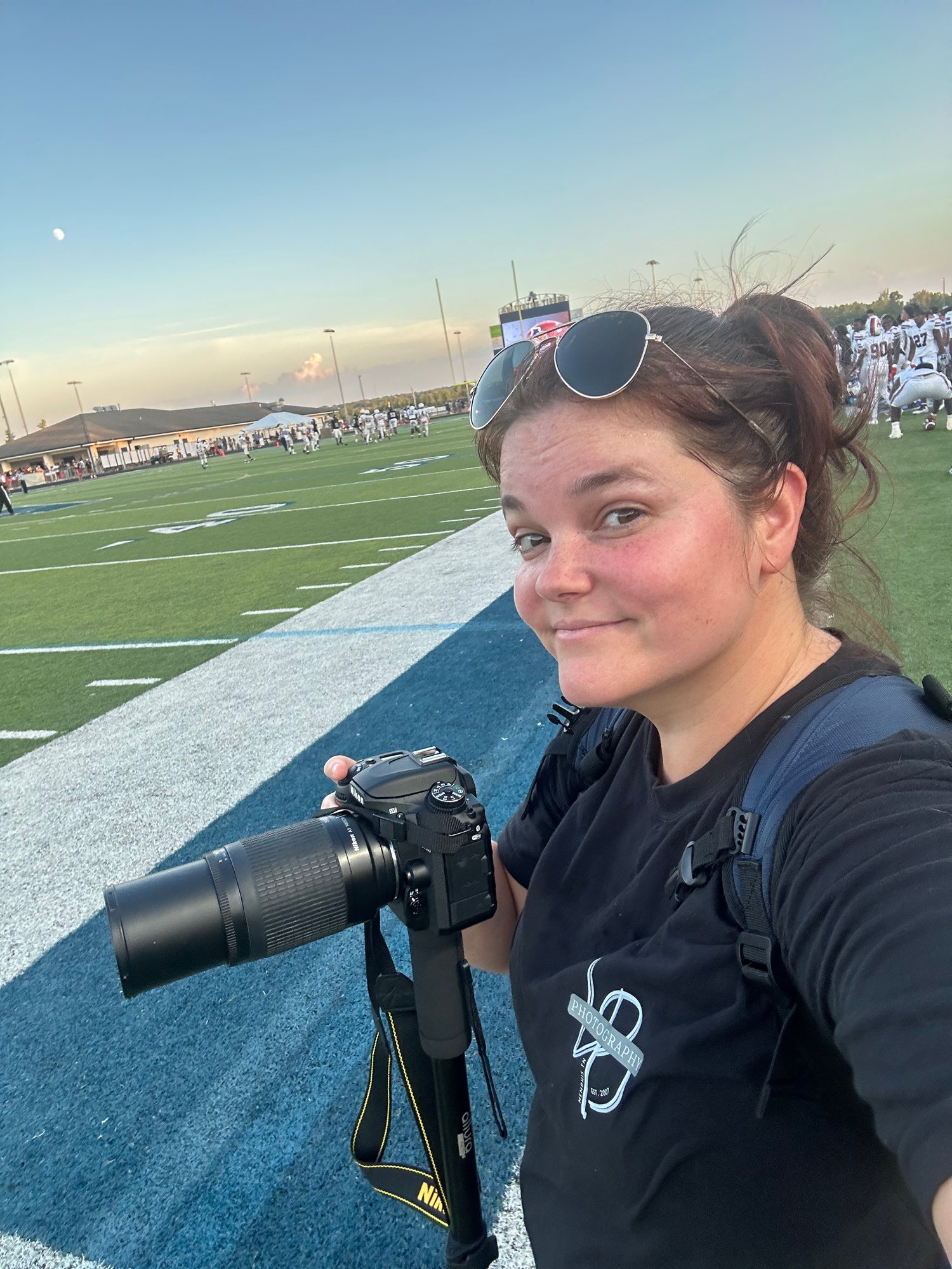 Young woman photographer holding a DSLR camera with telephoto lens at a football field during golden hour.