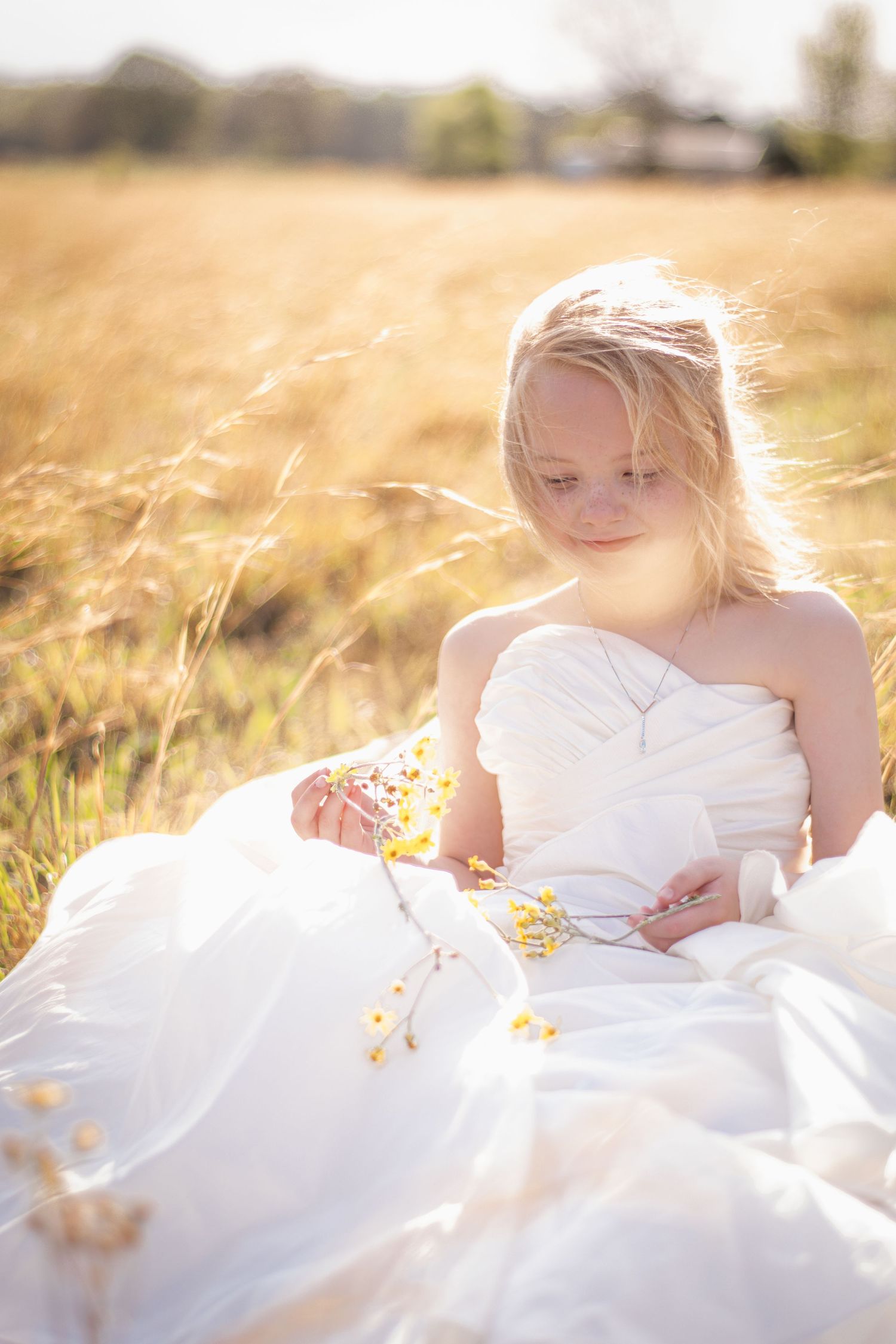 Young girl in white dress sitting in golden field holding yellow wildflowers in warm sunlight.