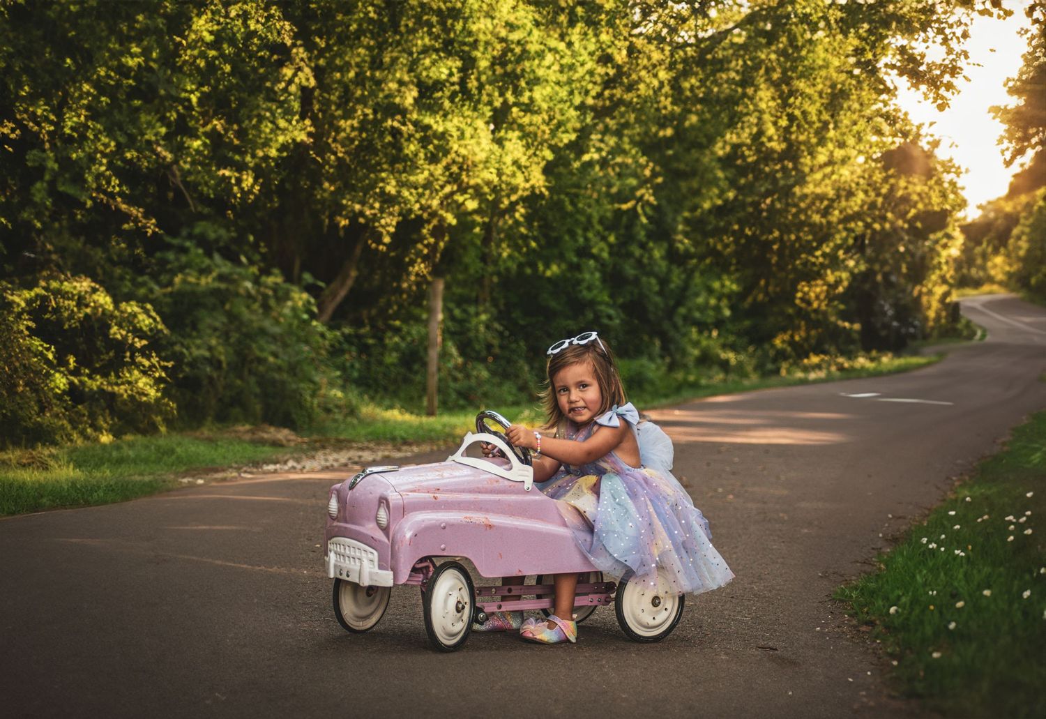 Young girl in blue dress drives pink vintage pedal car on sunlit road surrounded by lush green trees.