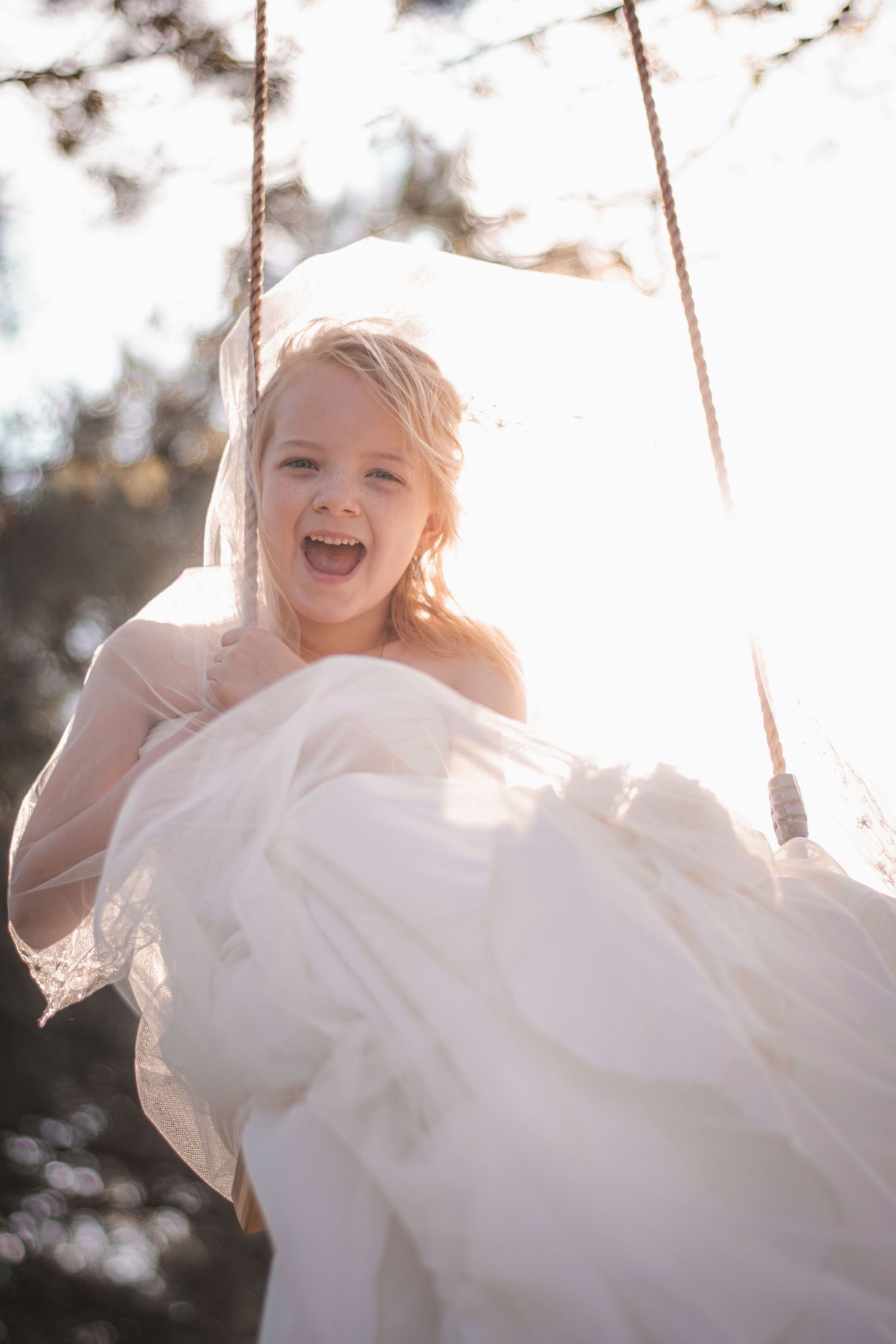 Joyful young girl in a white dress on a rope swing, backlit by golden sunlight with trees in background.