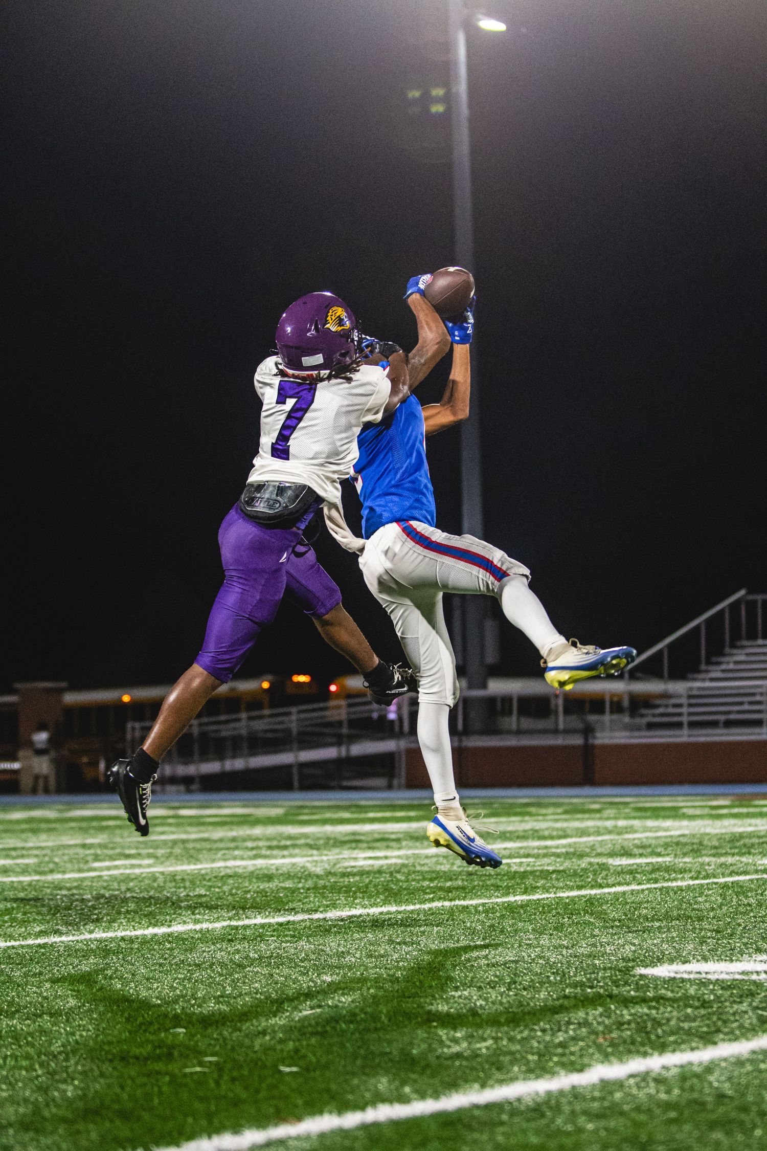 Two football players jump for the ball under stadium lights, number 7 in purple jersey competing against blue team player.