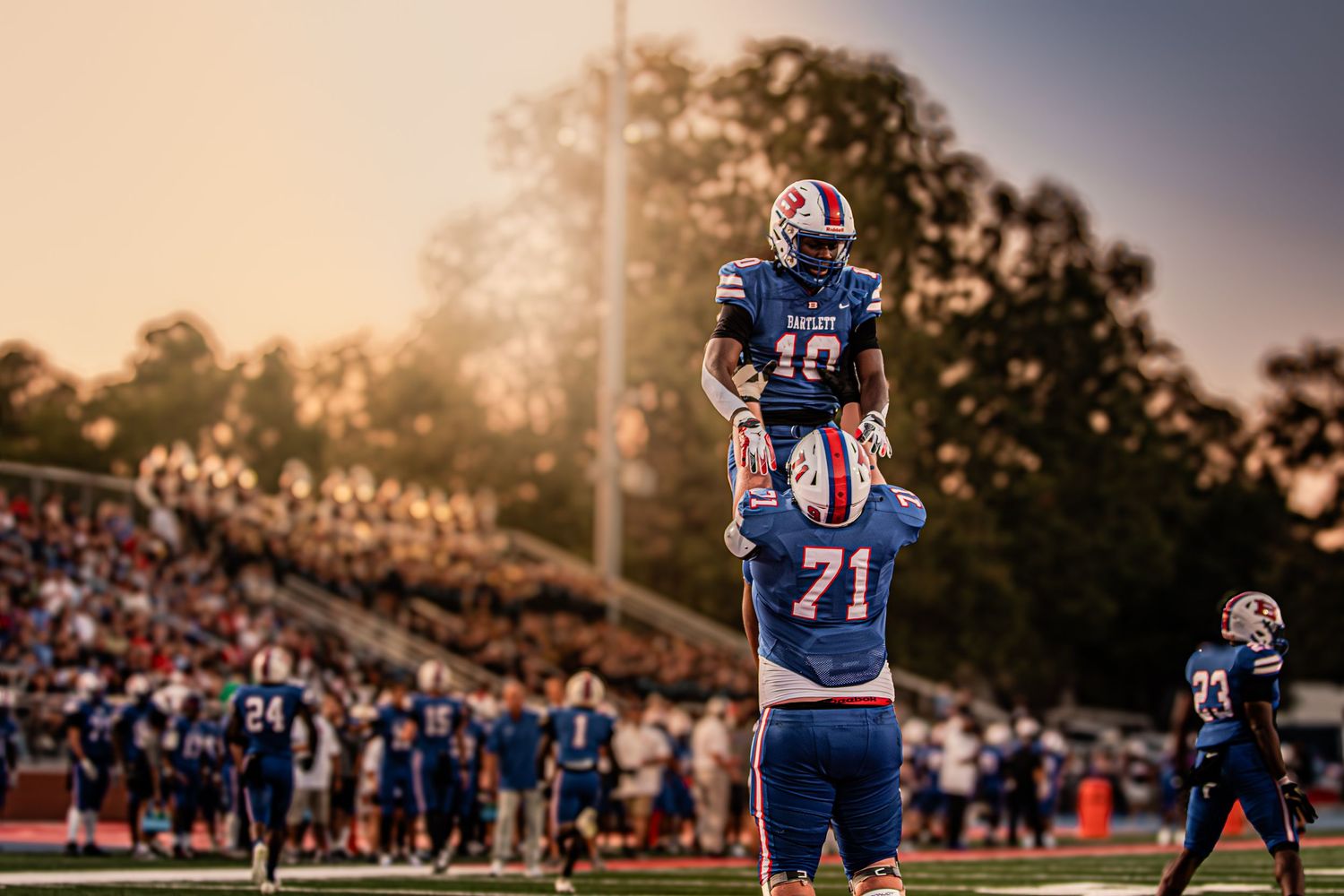 Two football players celebrate on the field at sunset with a crowded stadium in the background.