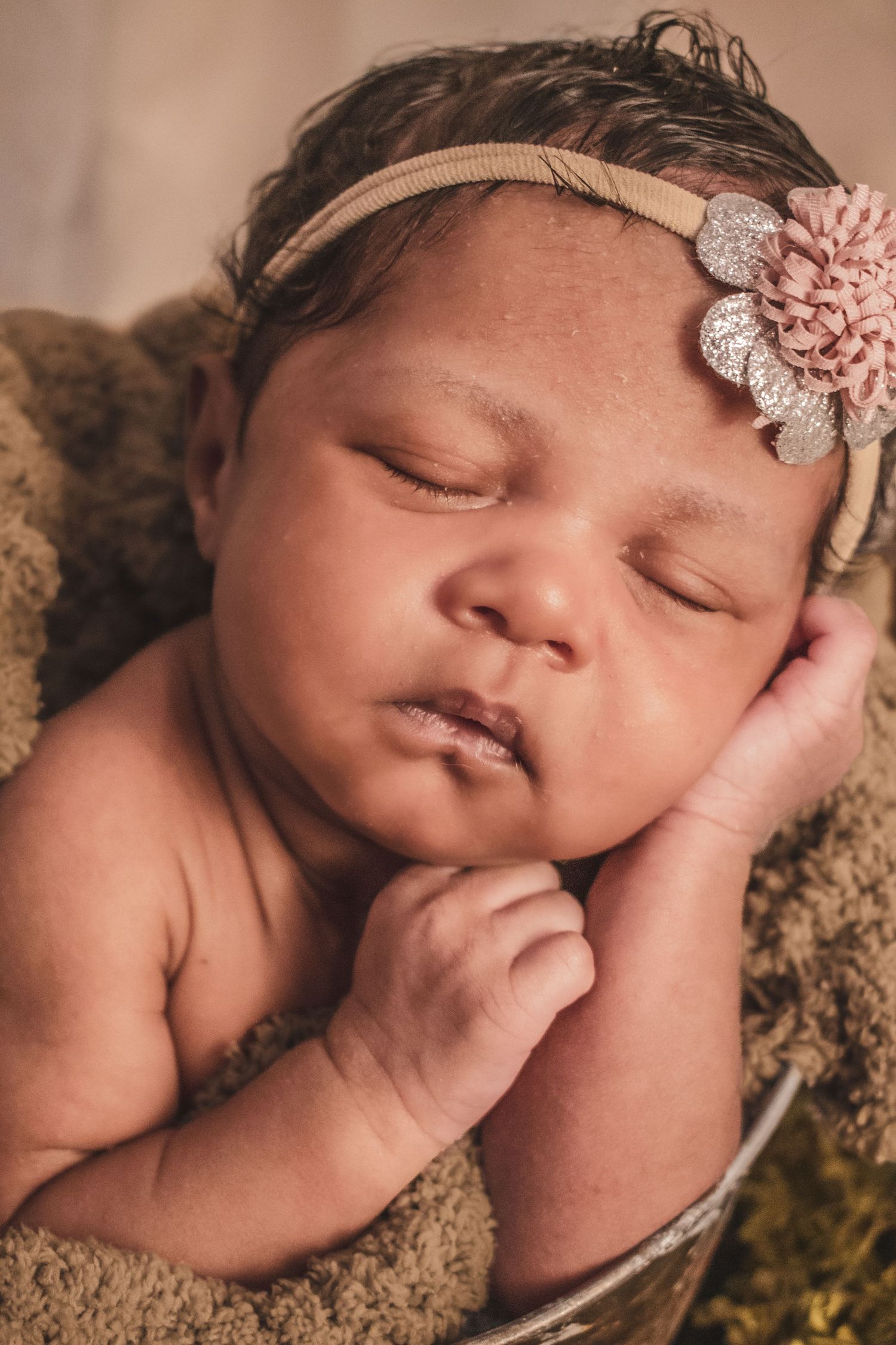 Sleeping newborn baby girl in a bucket wearing a pink floral headband, wrapped in soft brown blanket.