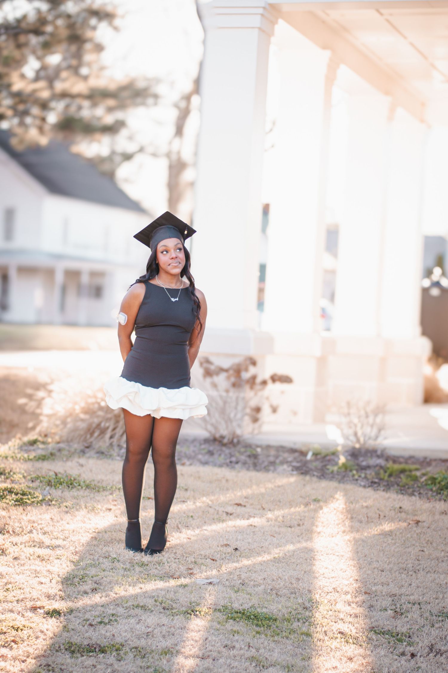 Young woman in graduation cap and black dress with white skirt poses outdoors near white columns on a sunny day.