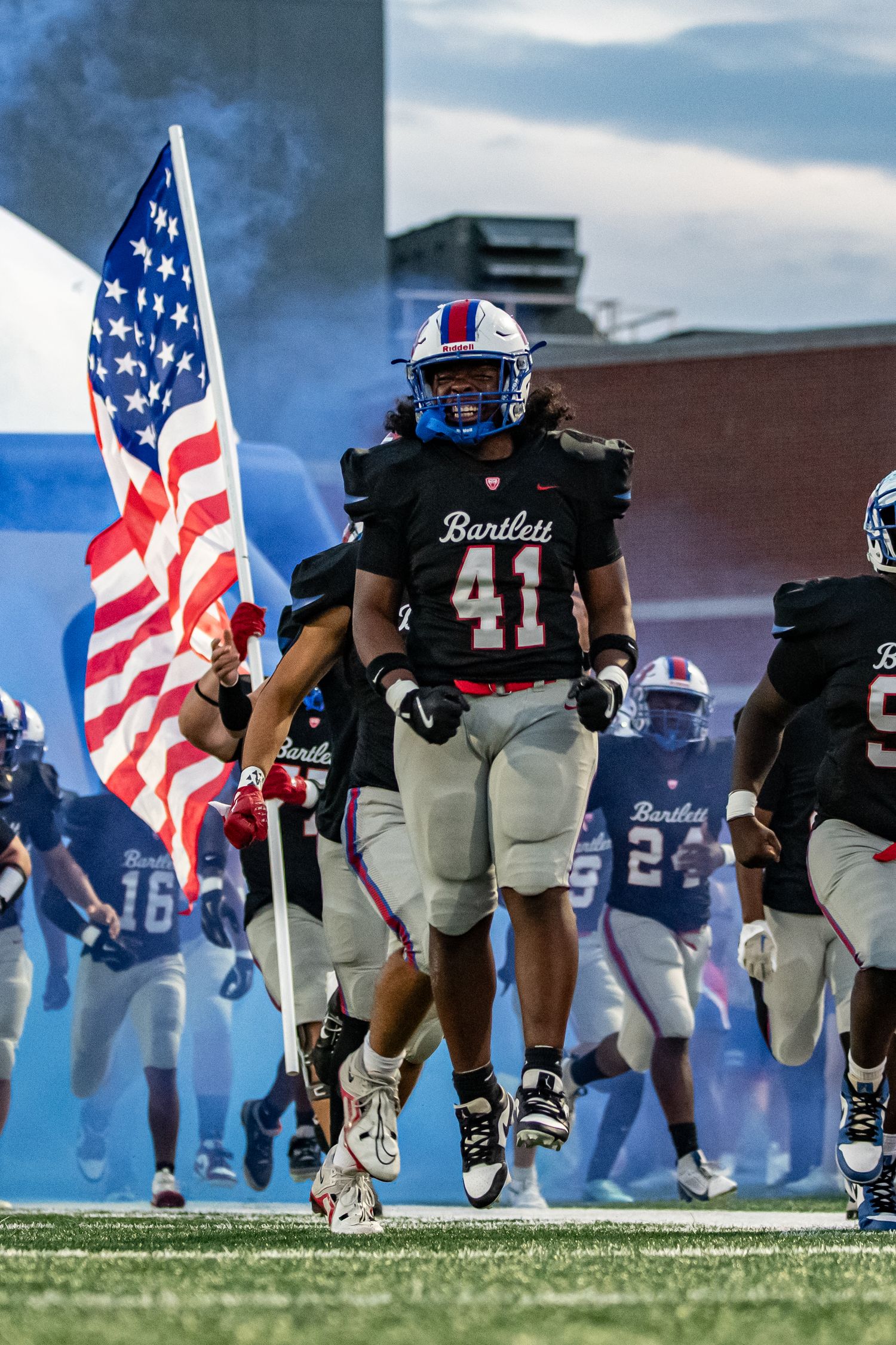 Bartlett football player #41 leads team onto field carrying American flag in black uniform amid blue smoke.
