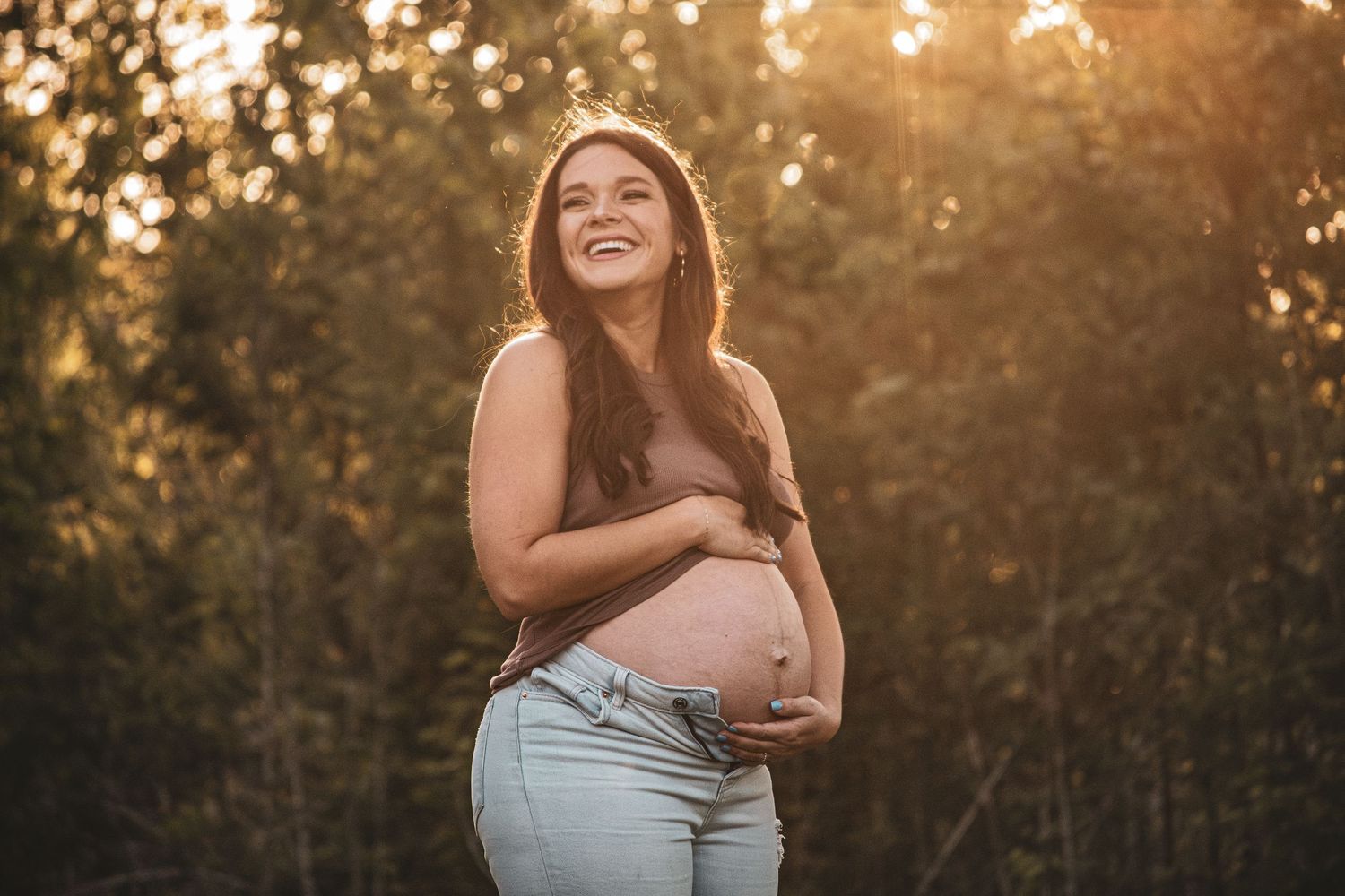 Smiling pregnant woman cradles baby bump in golden sunset forest setting.