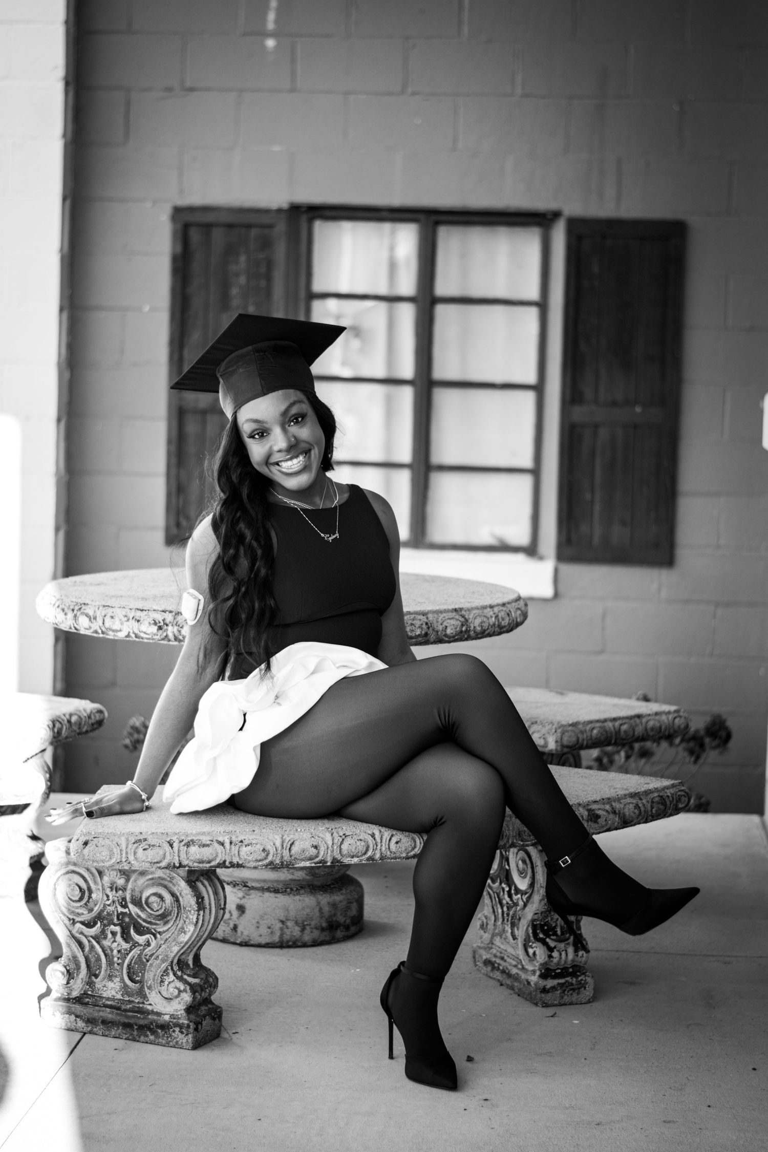 Black and white portrait of smiling graduate in cap sitting on ornate stone bench wearing heels.