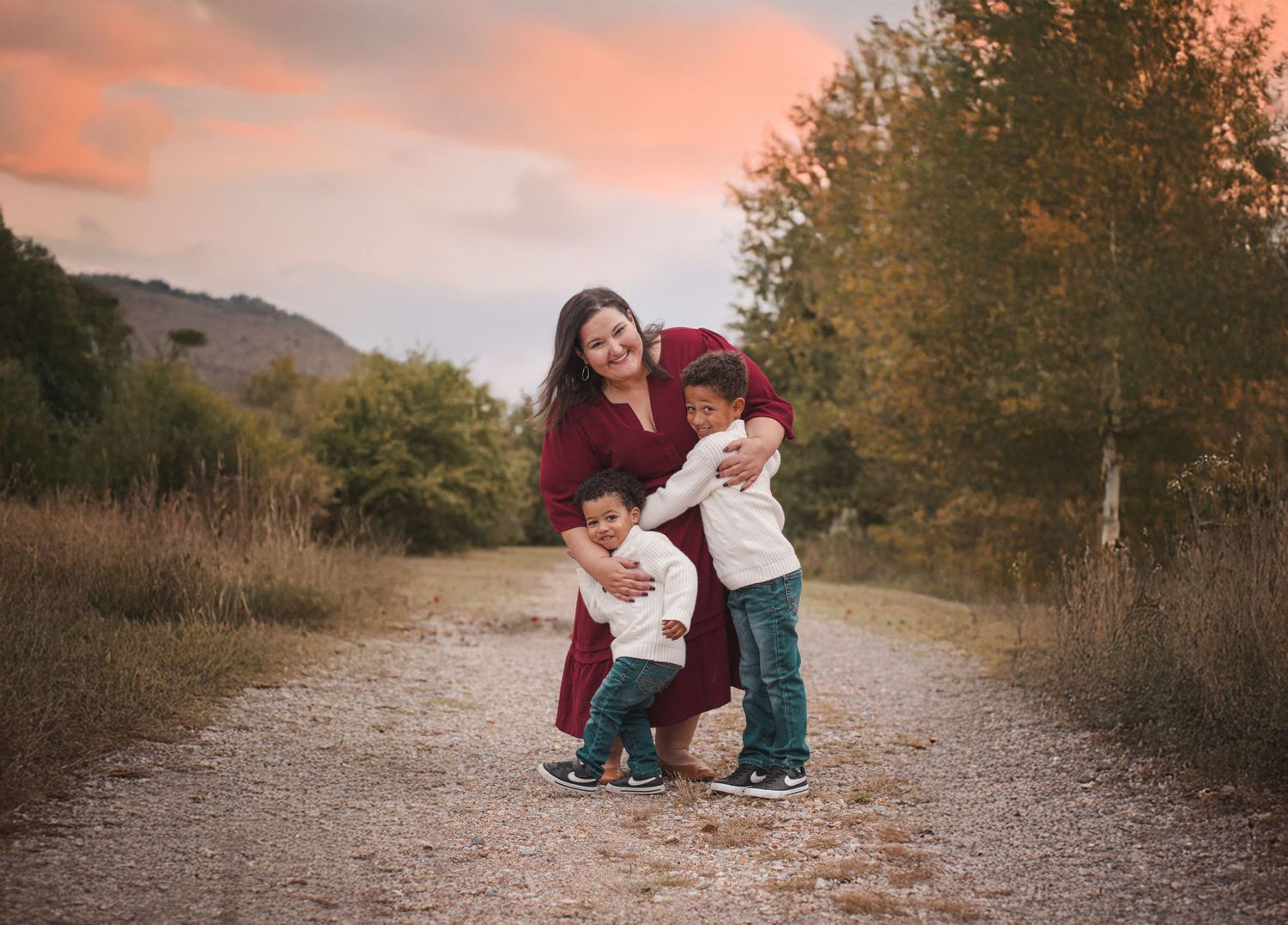Mother smiling with two young children on a gravel path during golden hour outdoors.