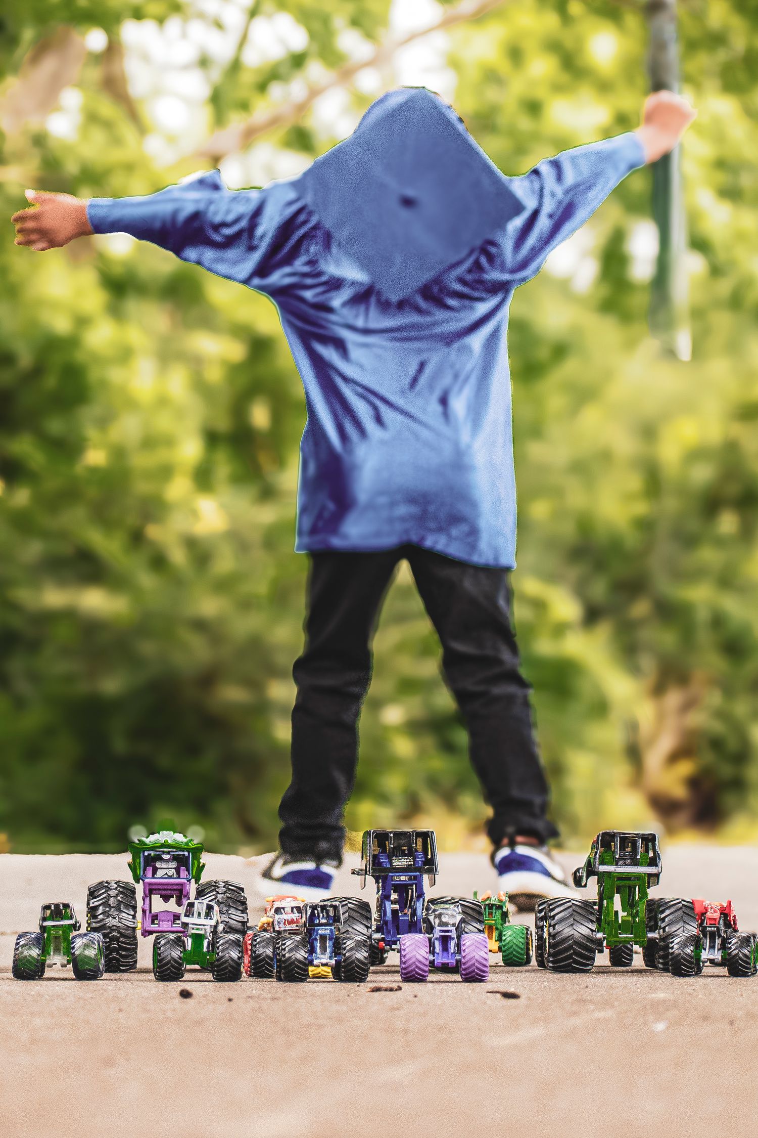Young boy in blue graduation gown stands with arms outstretched beside row of colorful toy trucks.