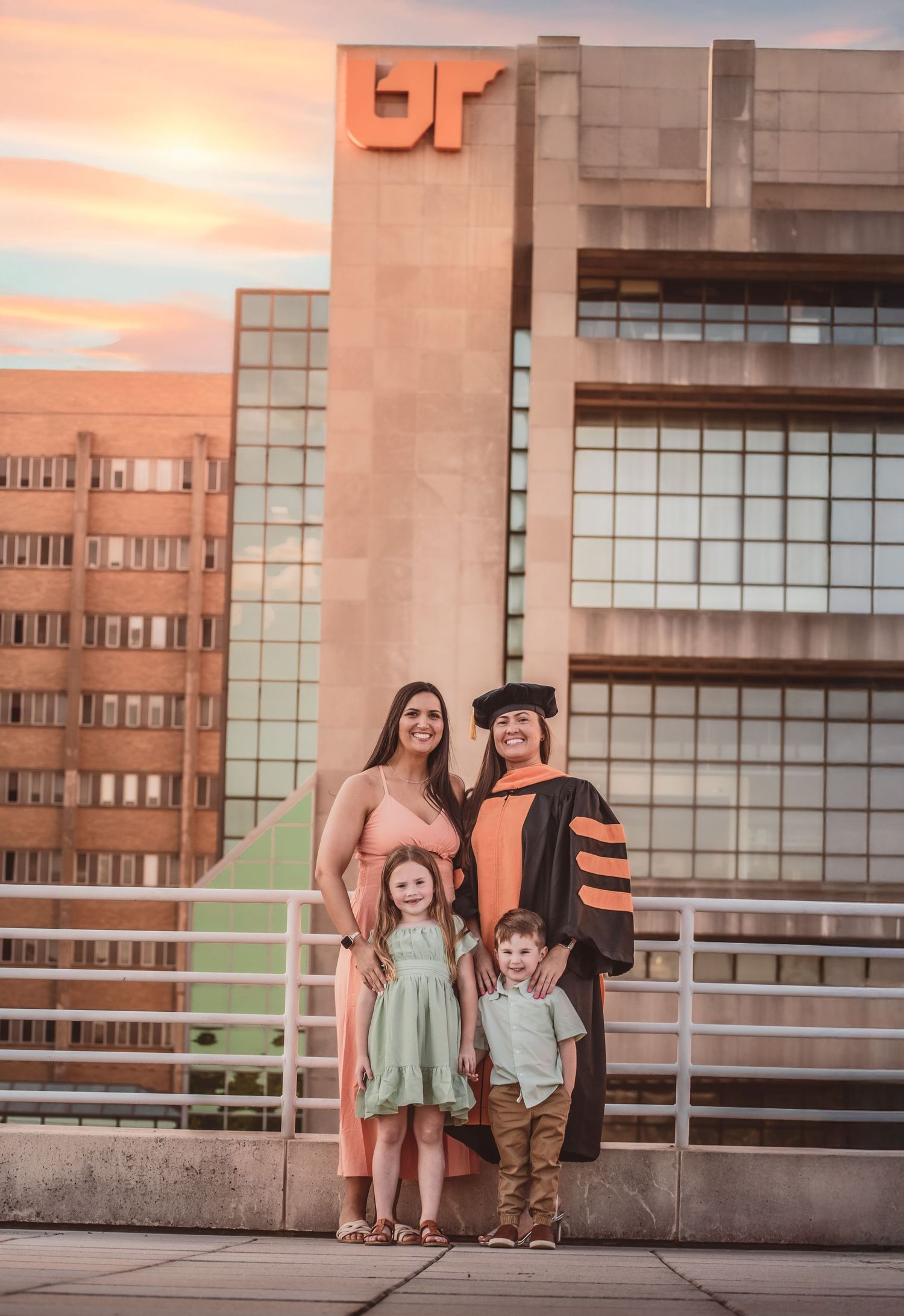 Graduate in doctoral regalia poses with family in front of UT building at golden sunset.