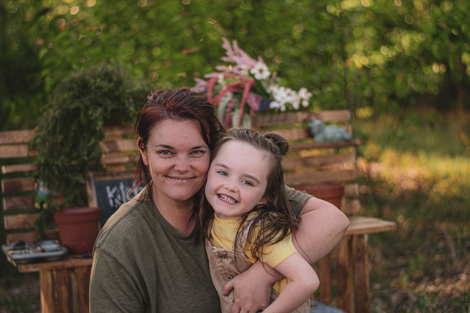 Smiling woman hugs young girl outdoors near rustic wooden benches with pink flowers in background.
