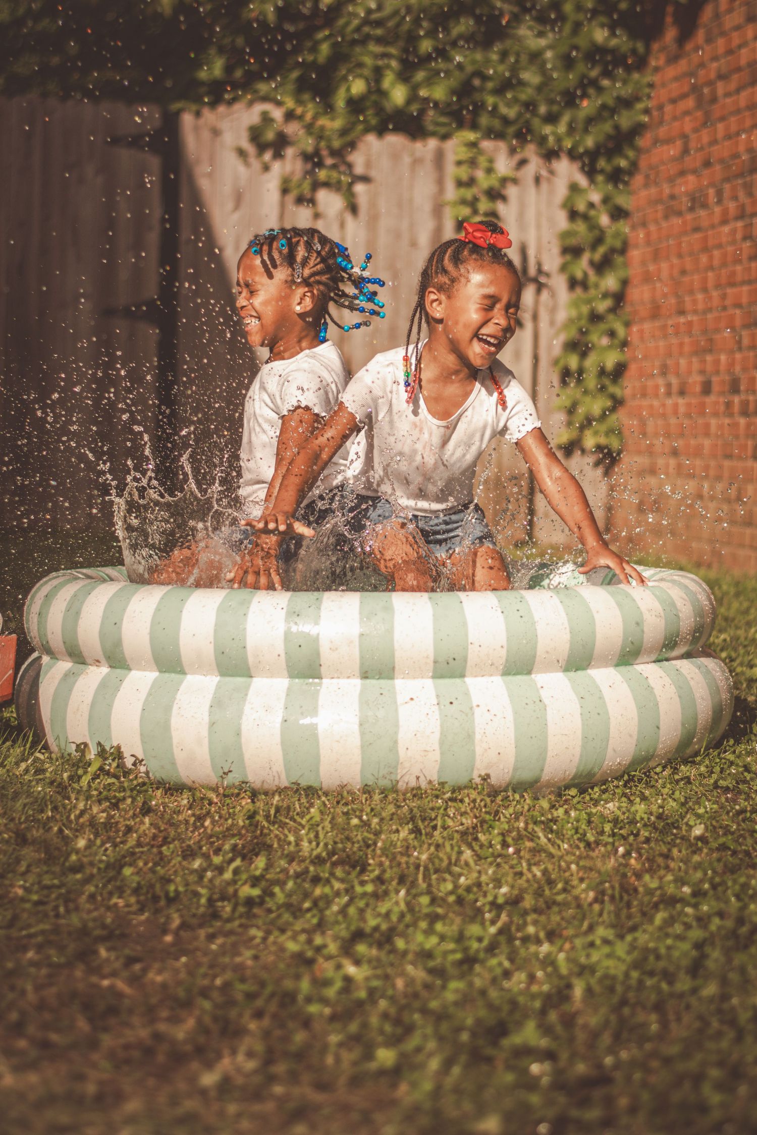 Two young girls laughing and splashing water in a striped inflatable pool in a sunny backyard. Lifestyle Photography