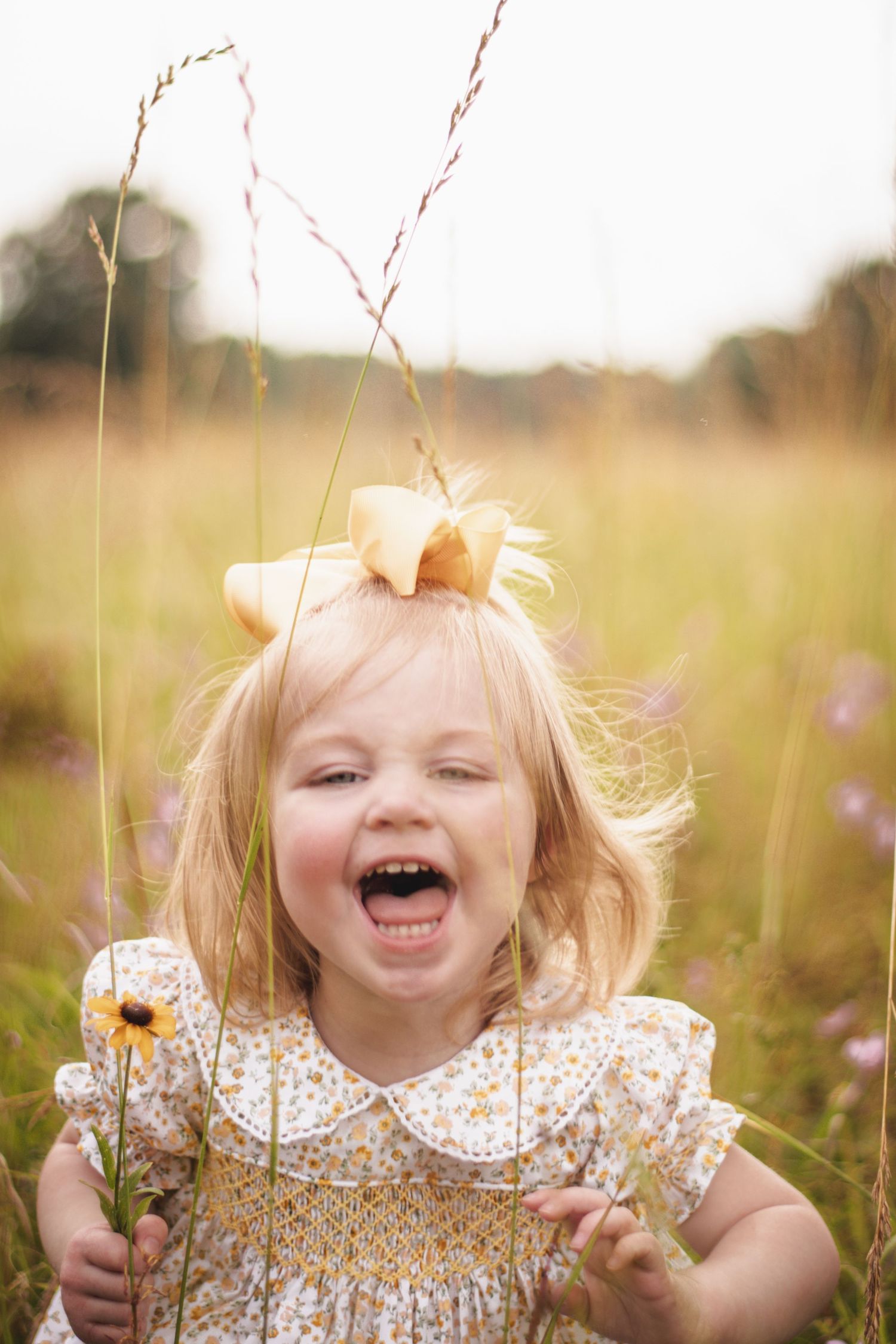 Joyful toddler girl with yellow bow laughing in golden meadow, wearing floral dress and holding wildflower.