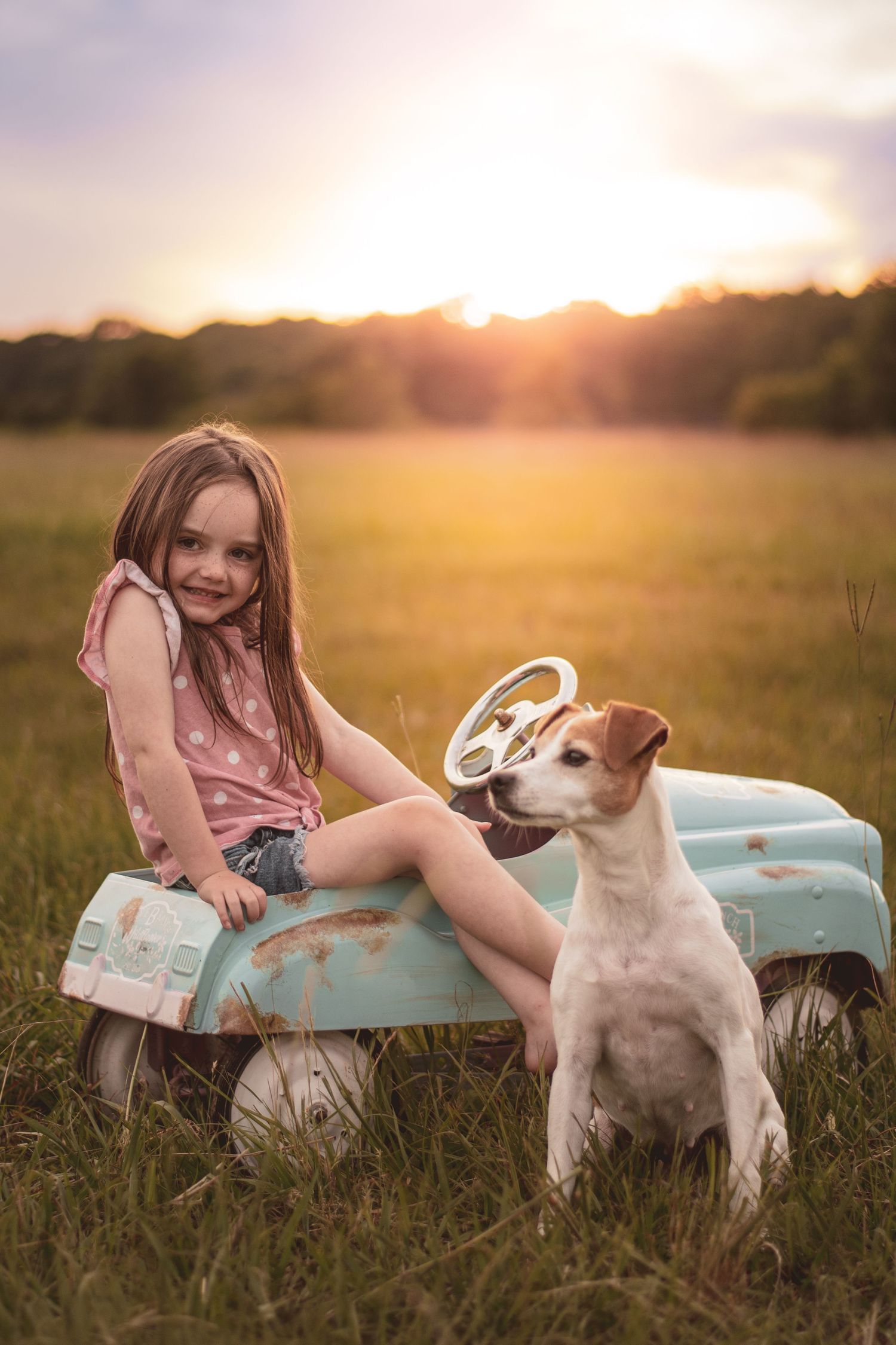 A smiling girl sits on a vintage toy car with a Jack Russell terrier dog in a sunny field at sunset.