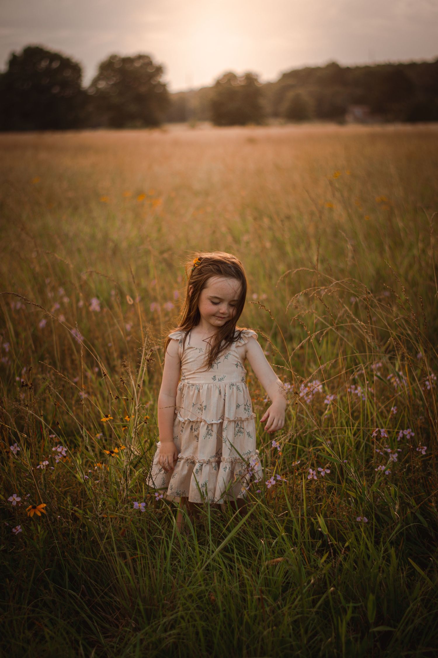Young girl in a white floral dress standing in a sunlit meadow with wildflowers at golden hour.