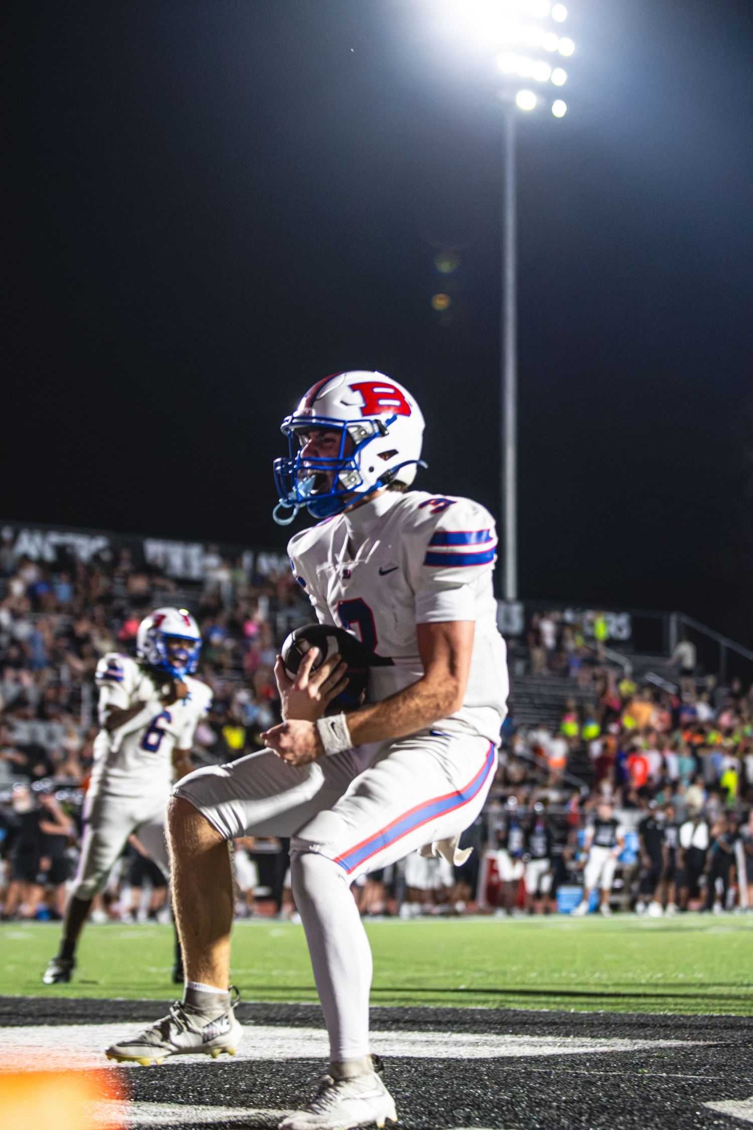 Football player in white and blue uniform carries the ball under bright stadium lights during a night game.