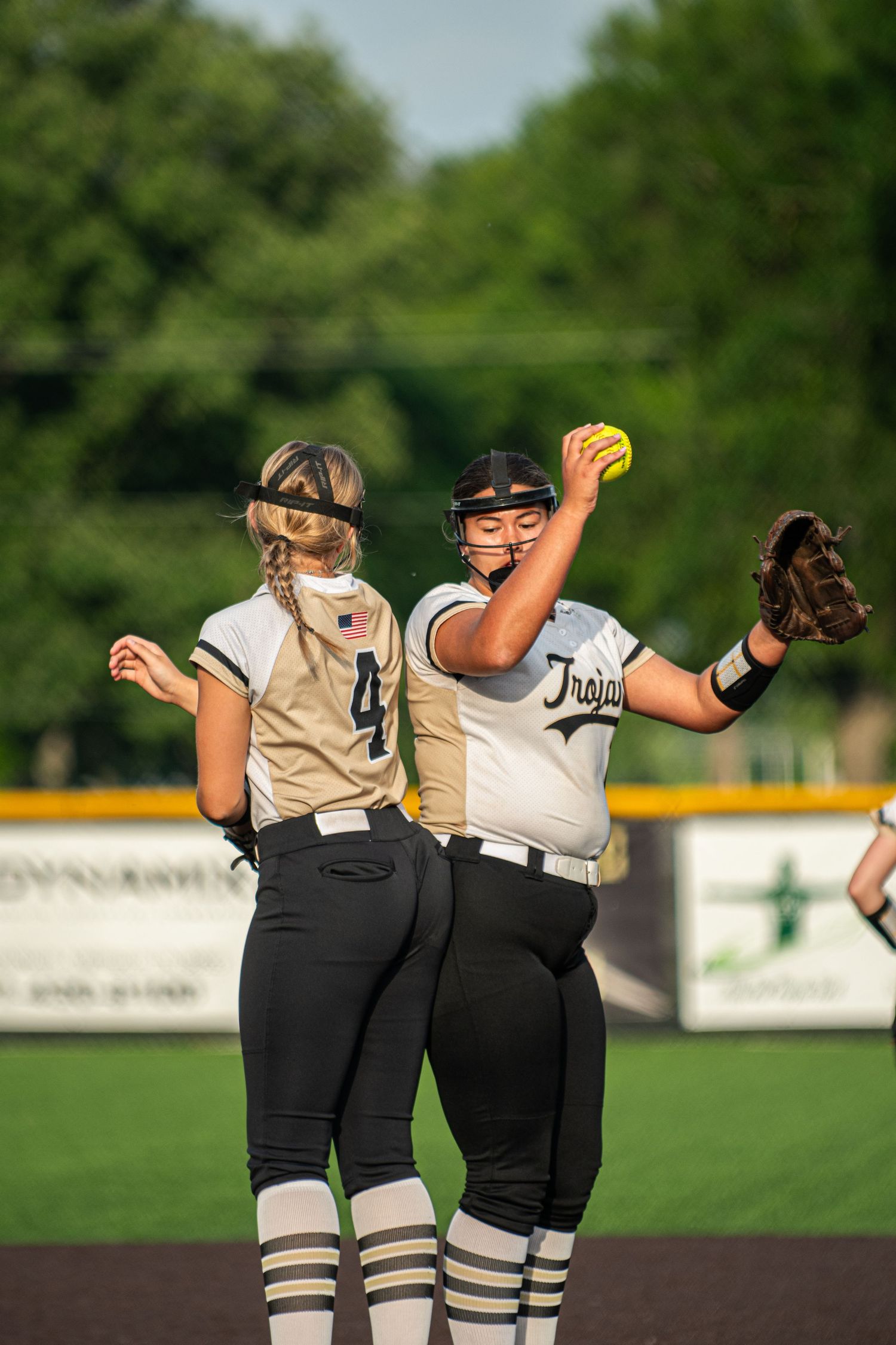 Two female softball players in Trojans uniforms on the field, one holding a yellow ball ready to pitch.