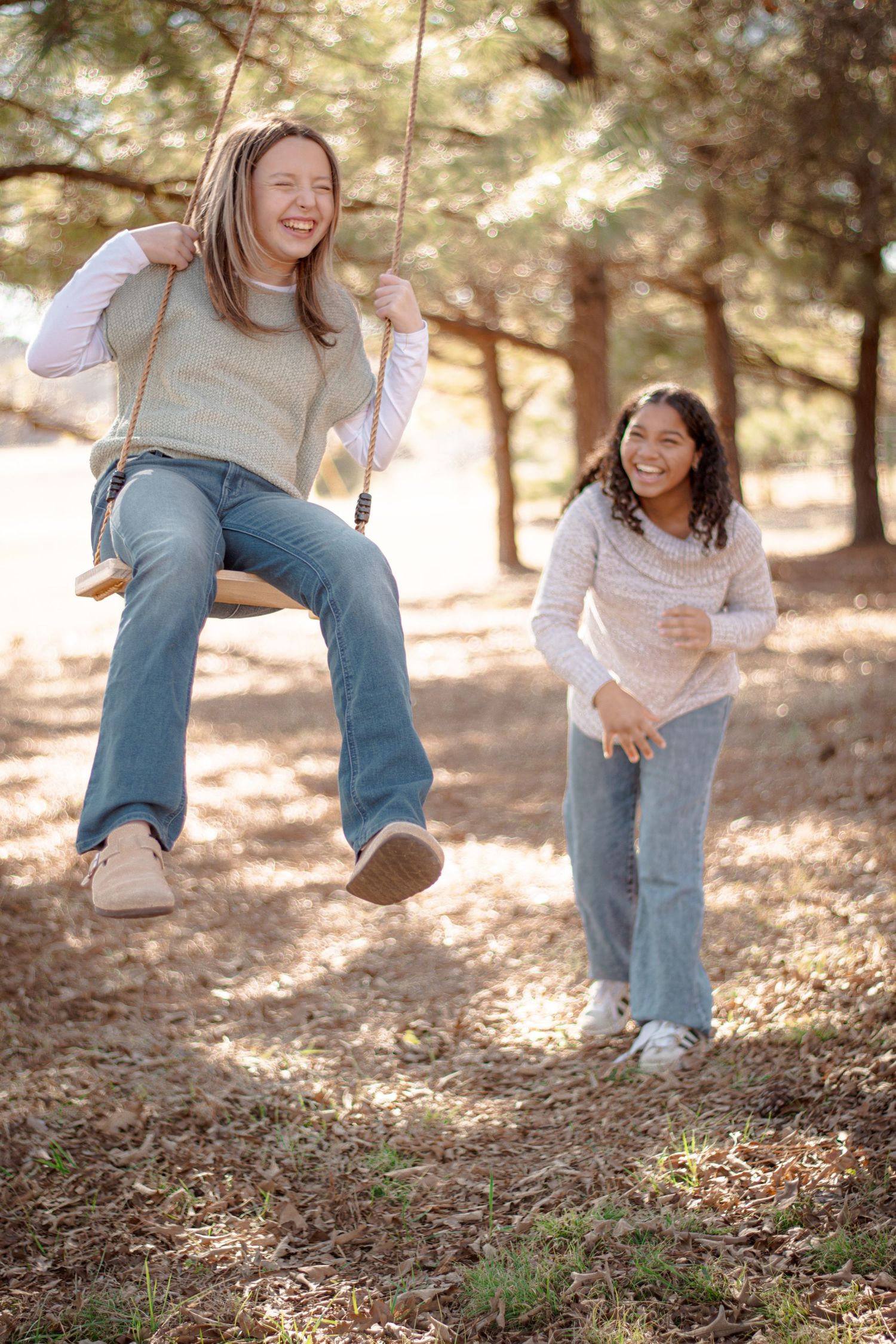 Two girls laughing outdoors, one on rope swing being pushed by friend among pine trees.