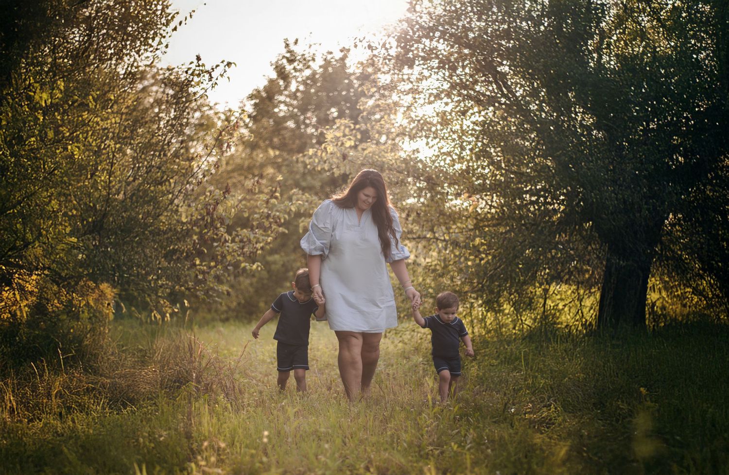 Mother in white dress walking with two young children through a sunlit forest meadow.
