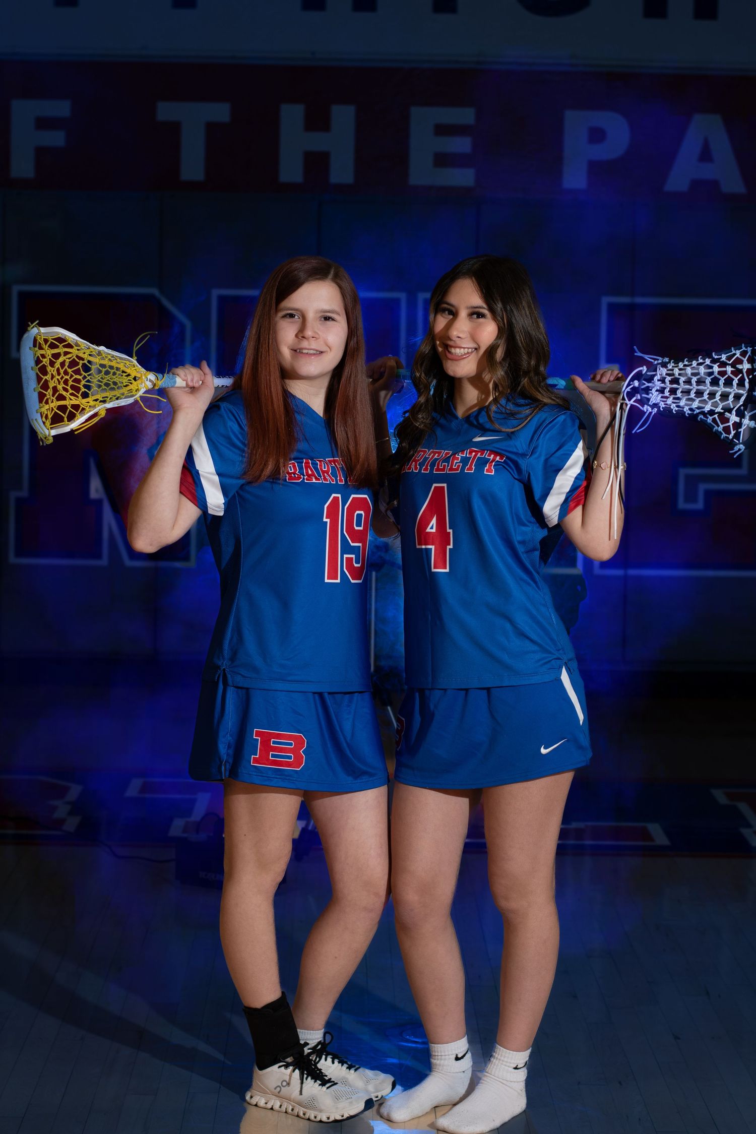 Two female lacrosse players in blue jerseys numbered 19 and 4 pose with lacrosse sticks during a team photo.
