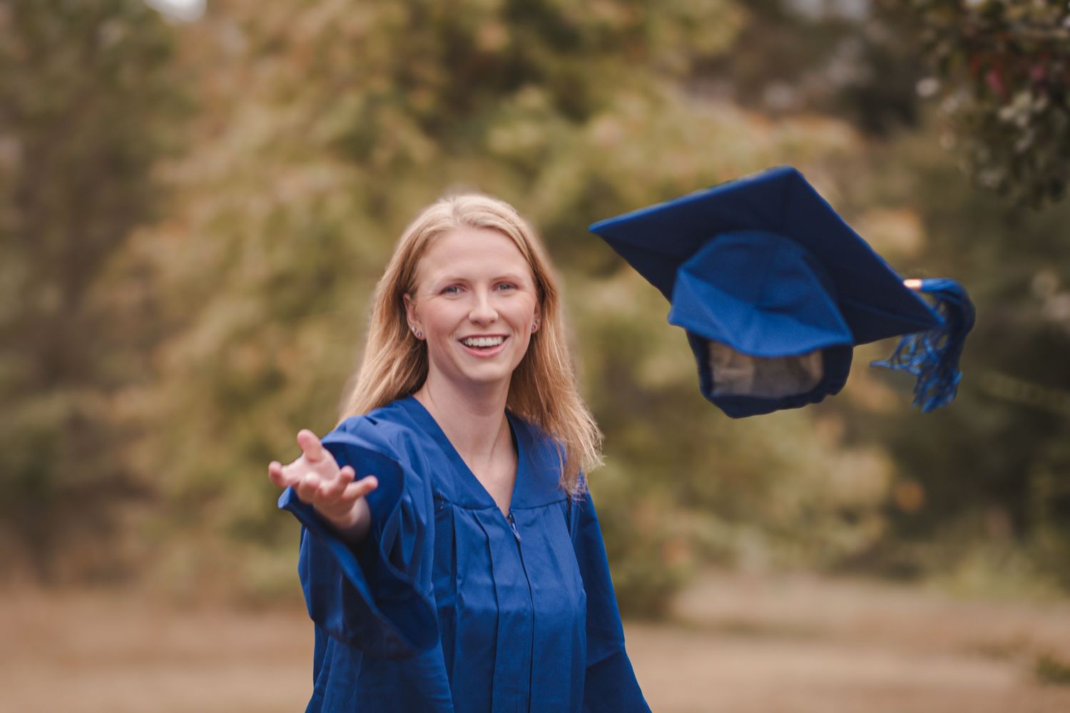 Smiling blonde woman in blue graduation gown tosses cap in the air outdoors among autumn trees.