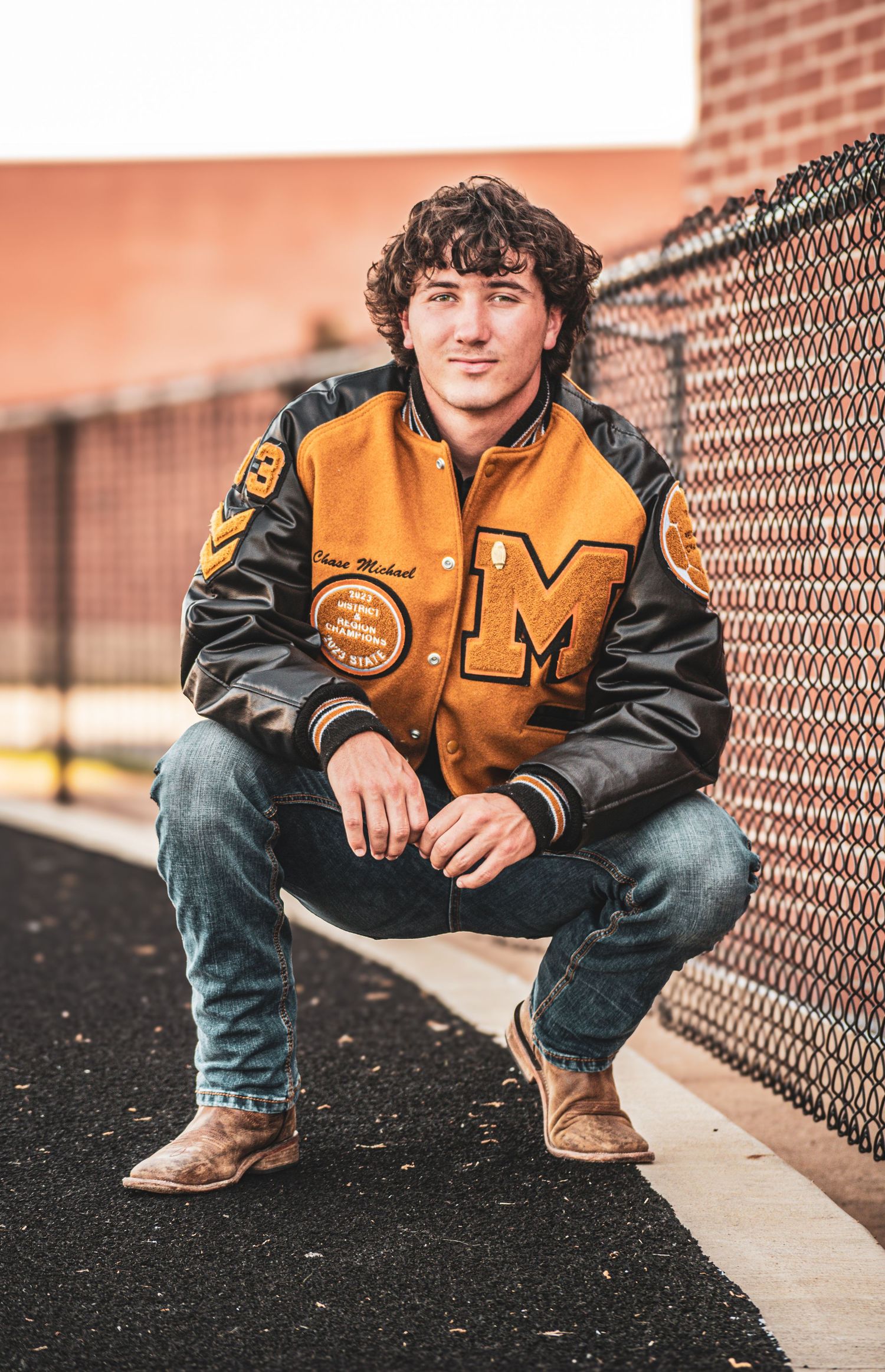 Teen boy in letterman jacket squats by chain-link fence for senior portrait.